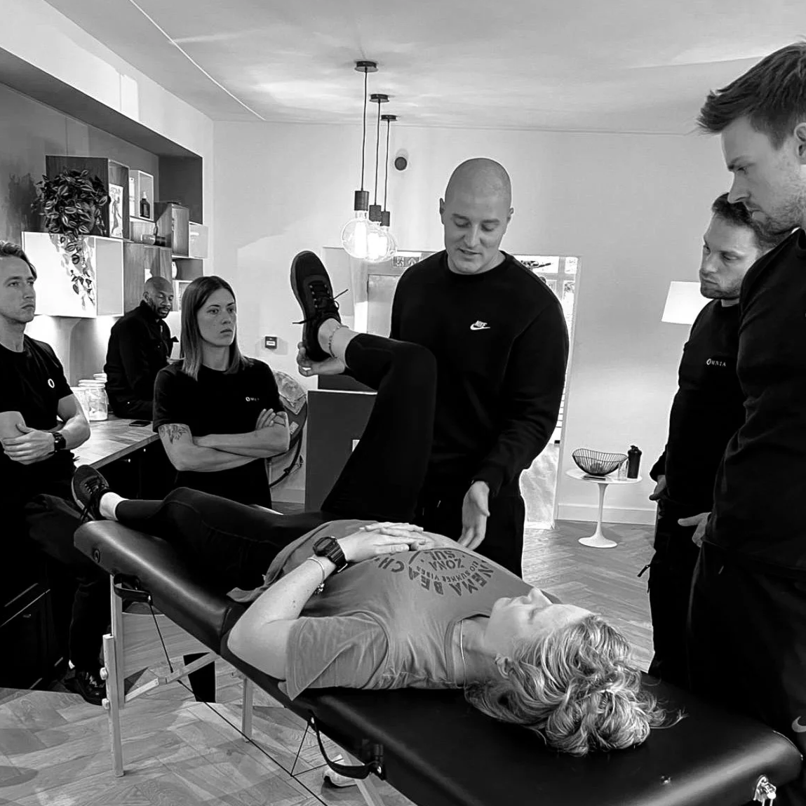 Group of people gathered around a woman lying on a therapy table, with a man demonstrating a physical exercise or therapy technique on her, in a room with modern decor and hanging lights.