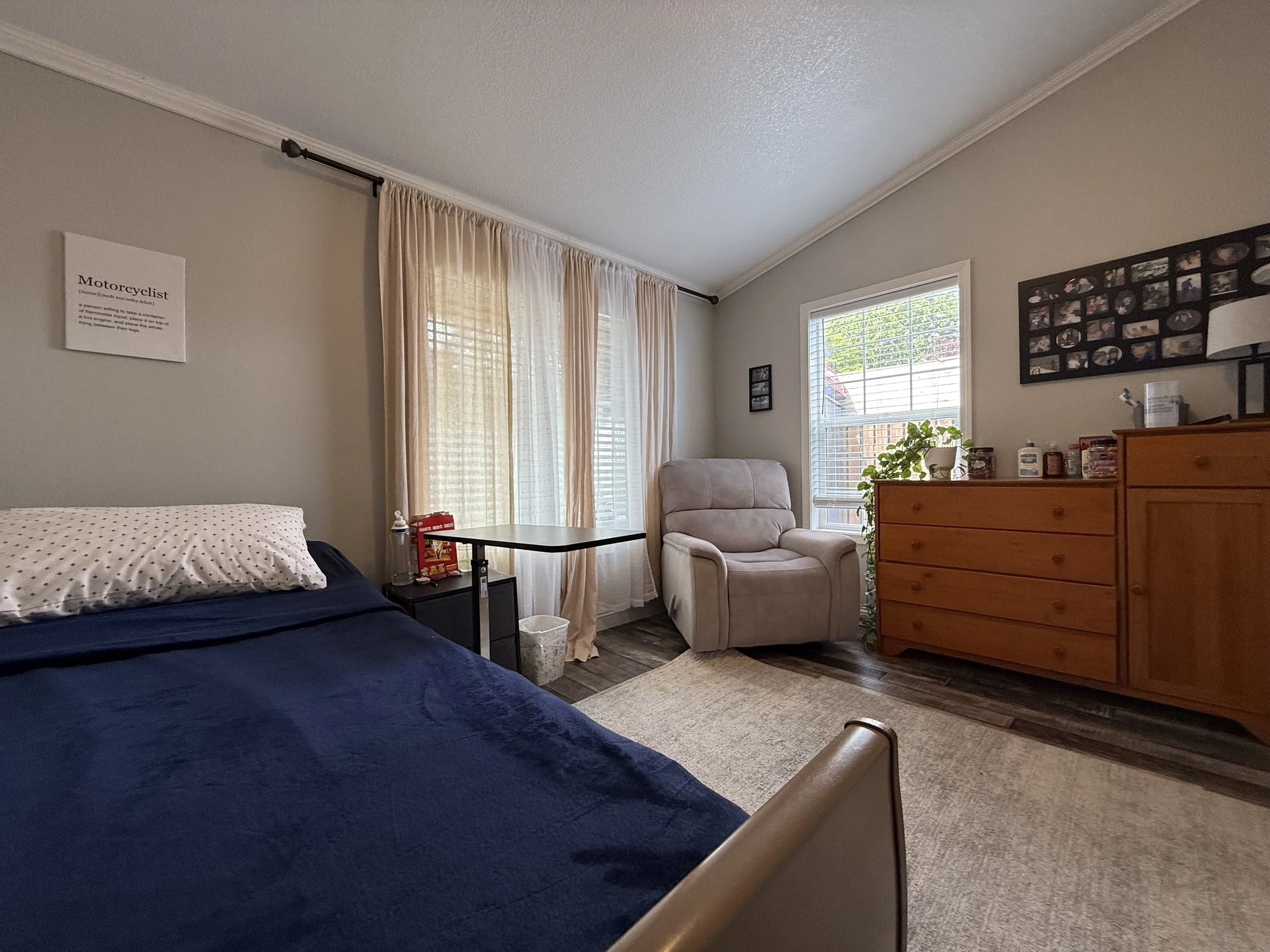 A bedroom with a bed in the foreground, beige curtains covering large windows, a beige armchair, a wooden dresser, and framed photos on the wall.