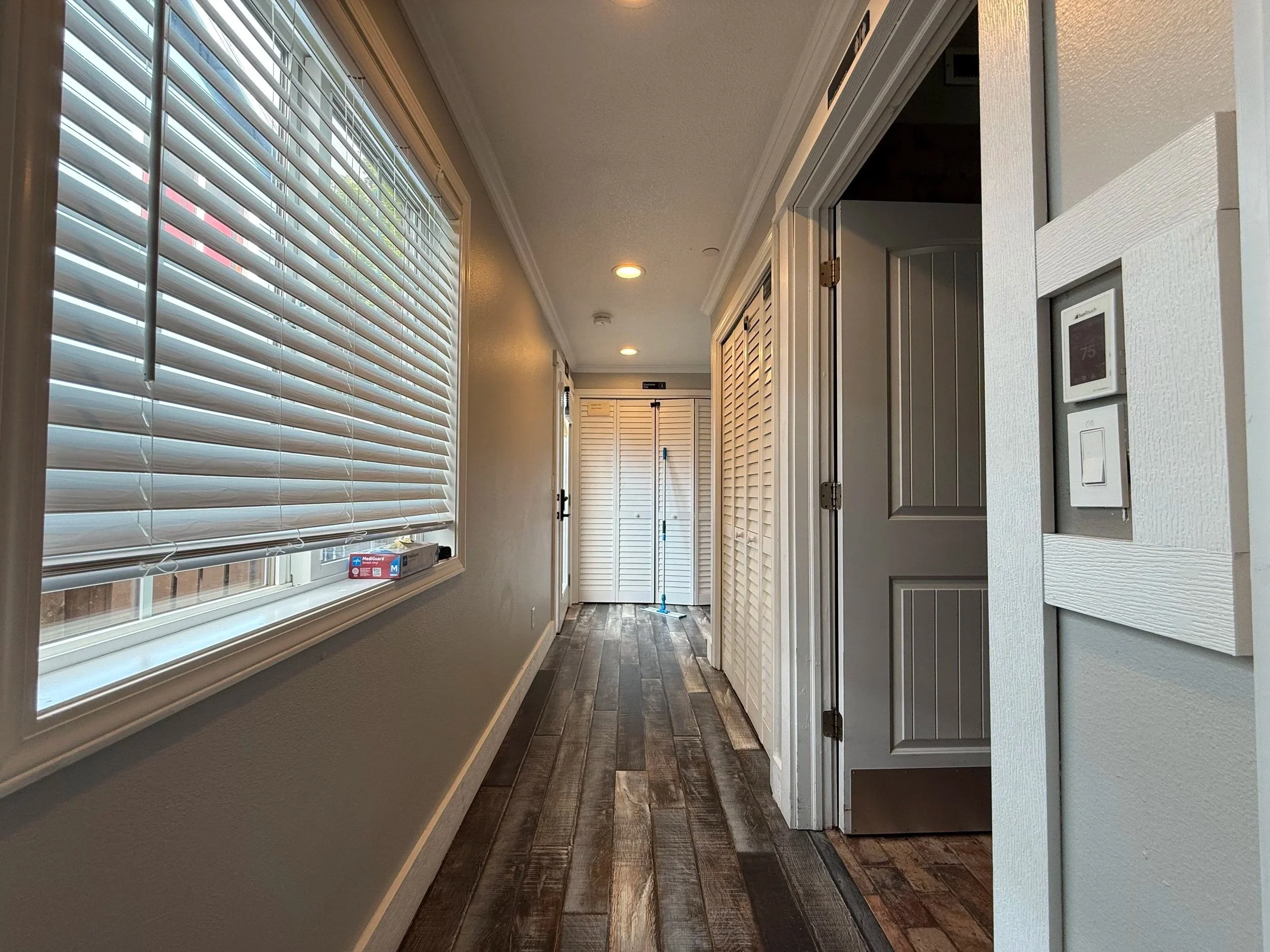 A hallway with large windows on the left side featuring white blinds, hardwood floors, ceiling lights, white walls, and various closed closet doors, with cleaning supplies visible at the end of the hallway.