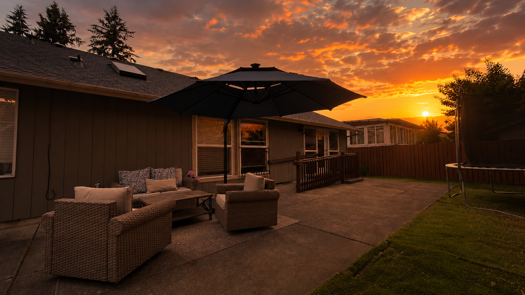 Sunset over a backyard patio with outdoor furniture, including wicker chairs, a cushioned sofa, a coffee table, and a large umbrella. There is a trampoline on the right and a wooden fence.