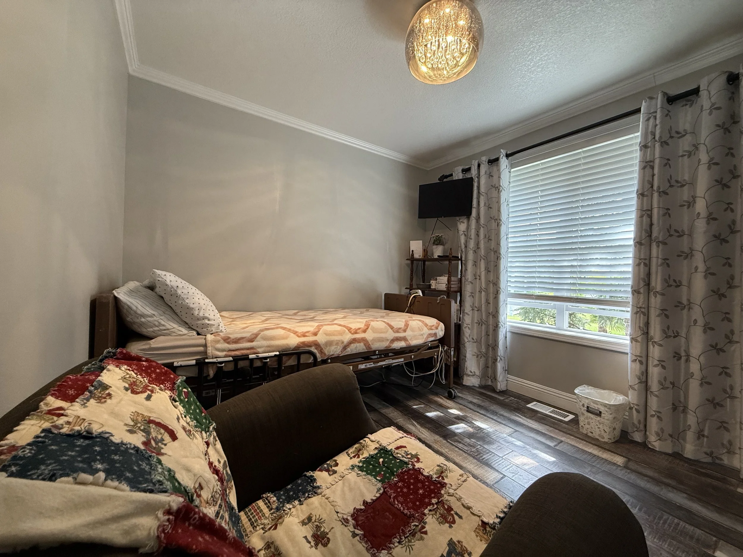A bedroom with a bed with Christmas-themed quilts, a wheelchair with a bedsheet, a window with curtains, a small TV, and a ceiling light.