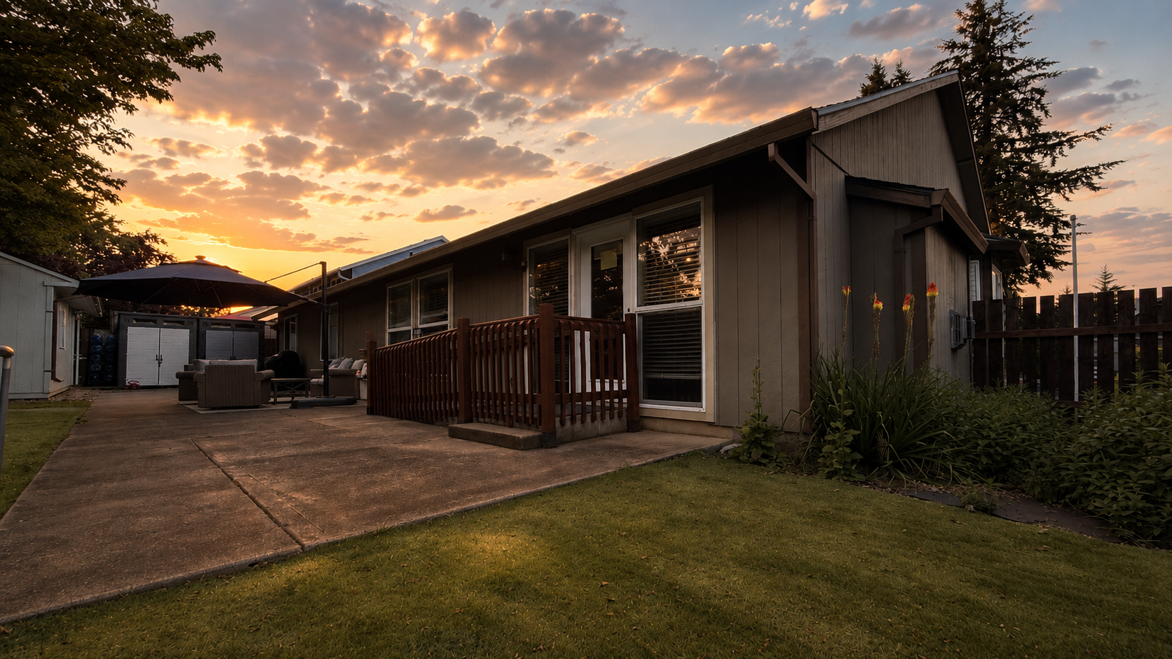 A backyard with a patio, outdoor furniture, a large umbrella, and a wooden deck, during sunset with a cloudy sky and a grassy lawn in the foreground.