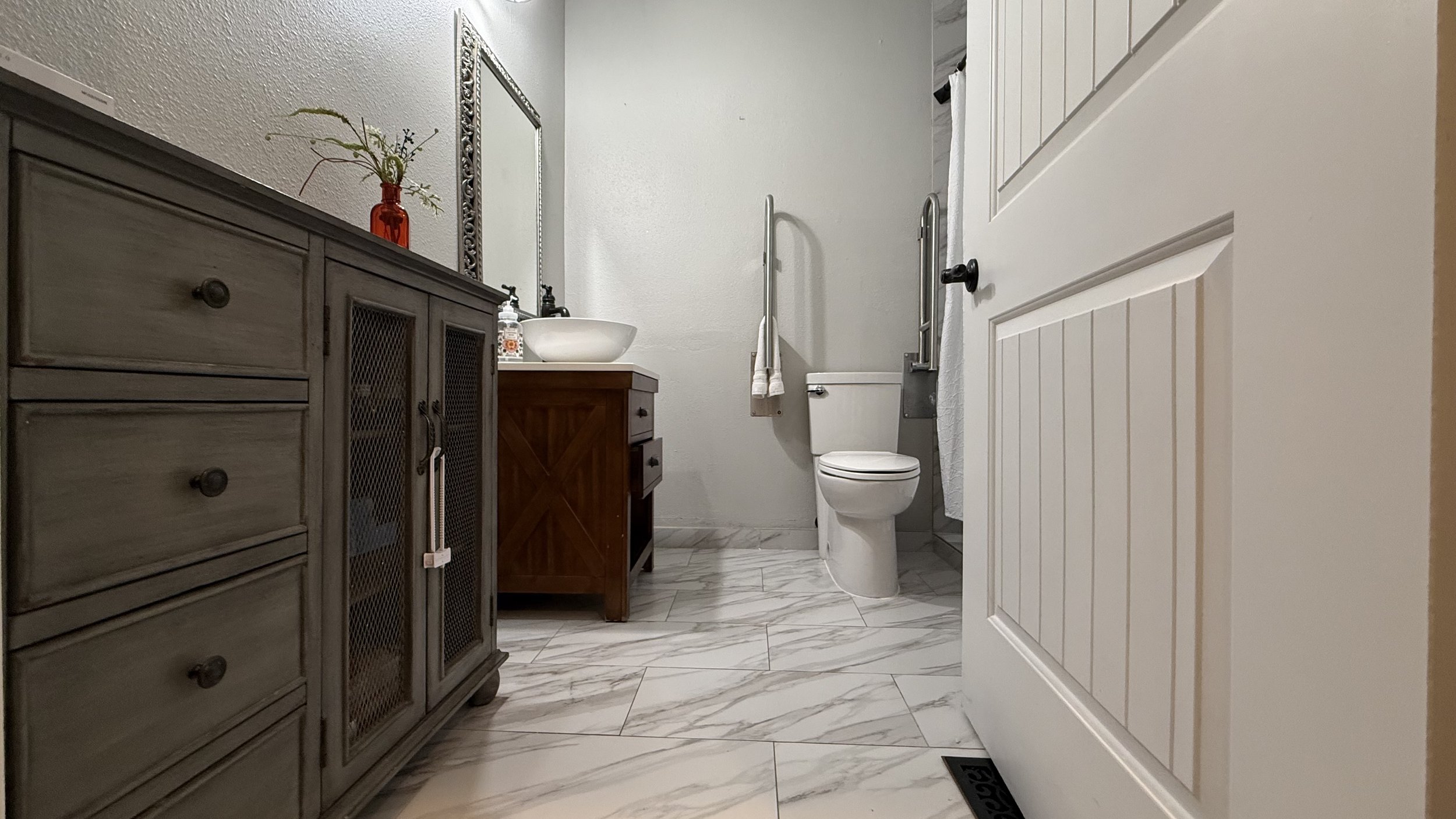 Bathroom with marble floor, gray textured wall, wooden vanity with a bowl sink, mirror, white toilet with grab bars, and a partially open white paneled door.