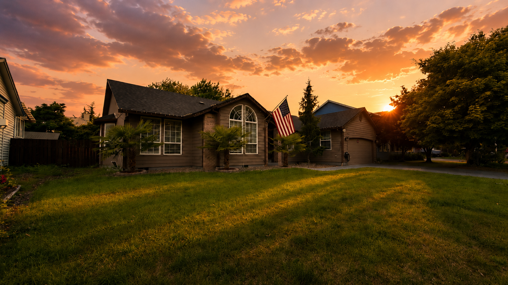 A suburban house during sunset with a well-kept lawn, palm trees in front, and an American flag hanging from the porch, surrounded by other houses and trees.