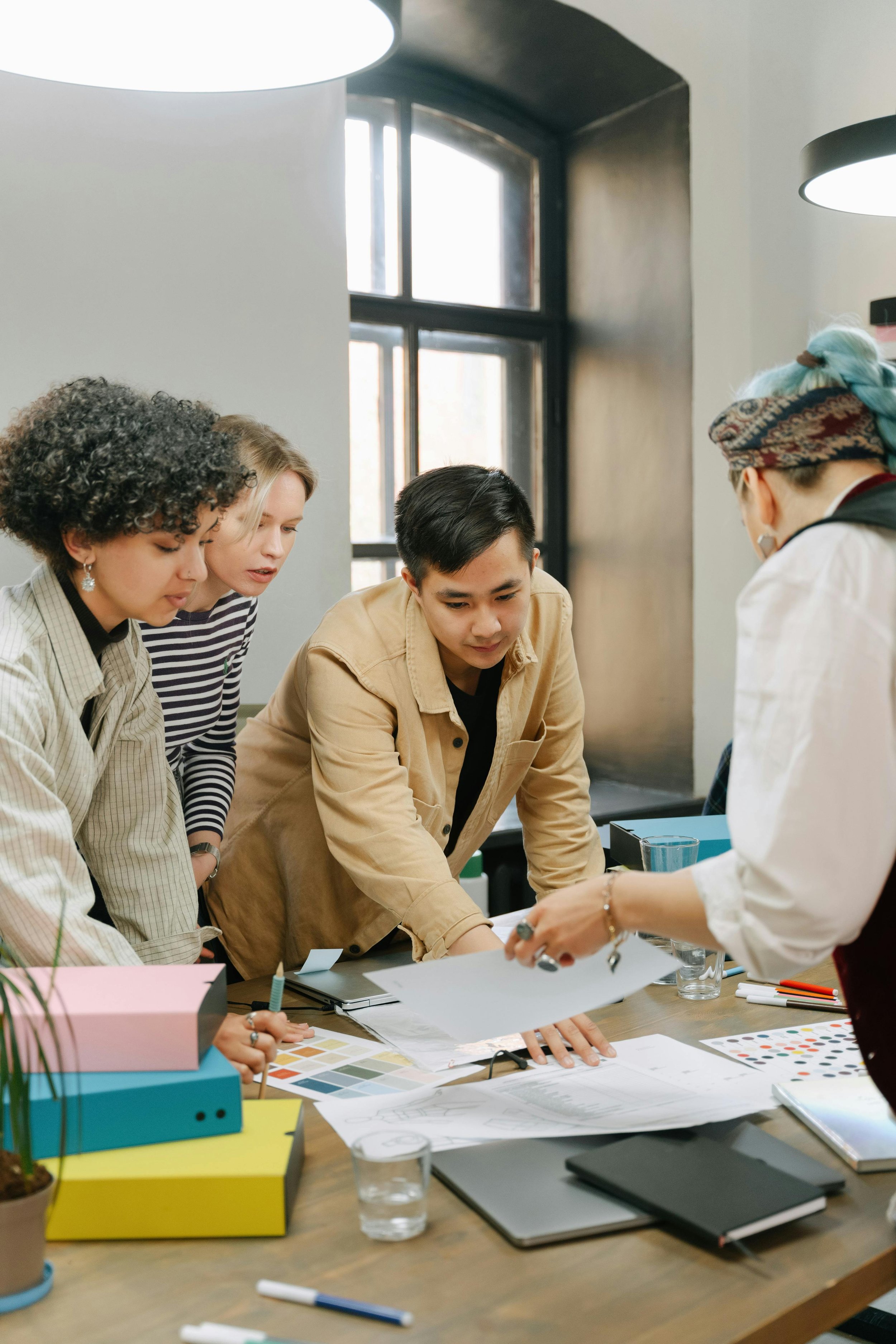 A diverse group of five people gathered around a table, looking at papers and design materials in a bright, modern office with large windows and hanging lights.