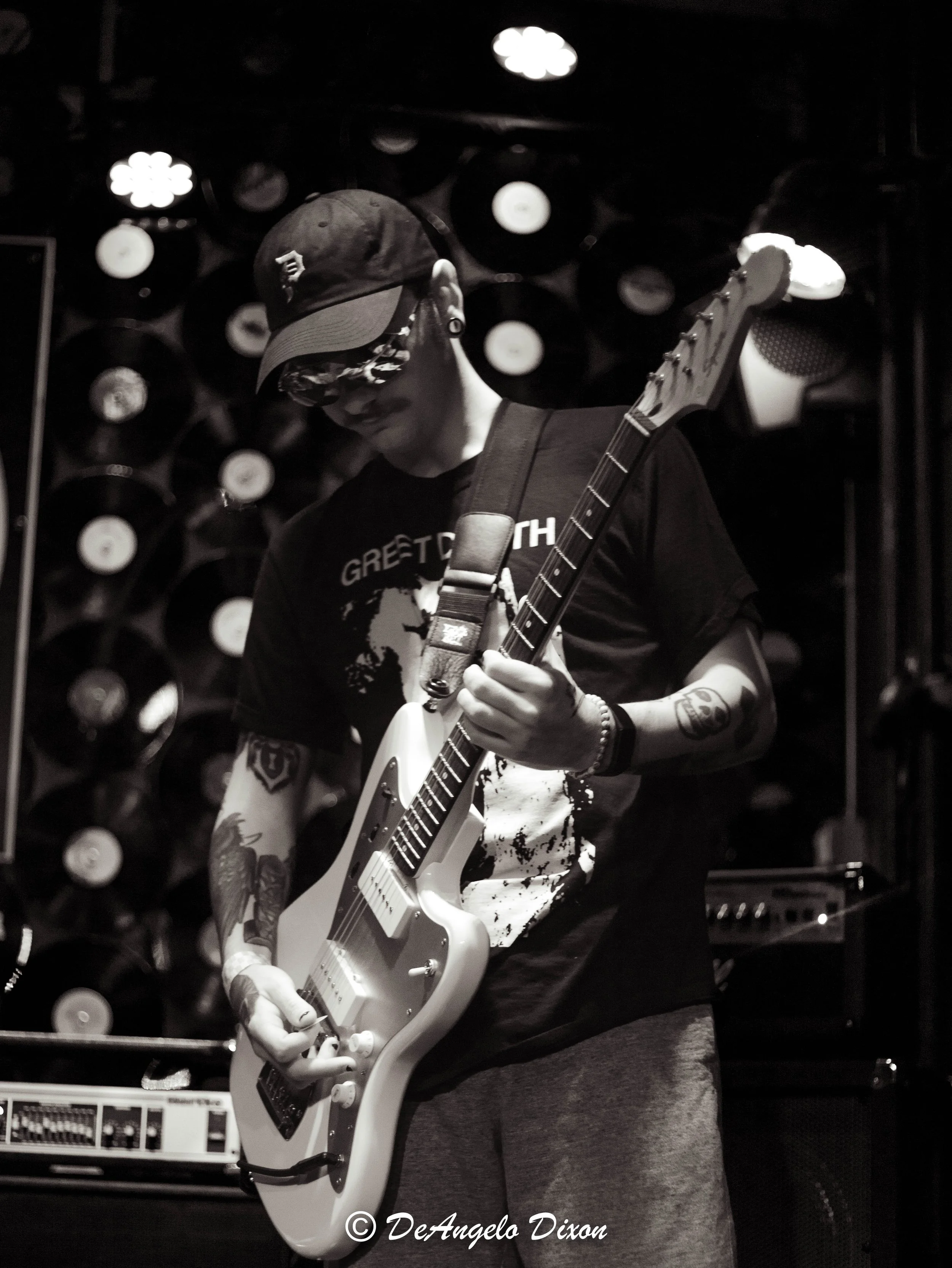 Person playing electric guitar in a dimly lit room with records on the wall in the background.