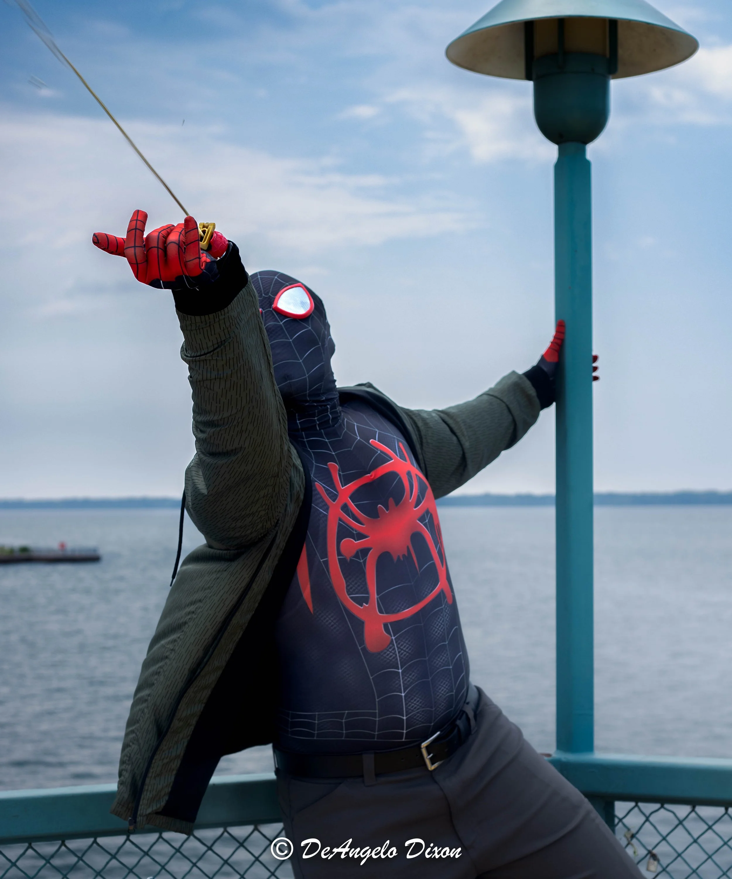 Person wearing Spider-Man themed clothing and gloves, holding onto a blue railing near a body of water, with a cloudy sky above.