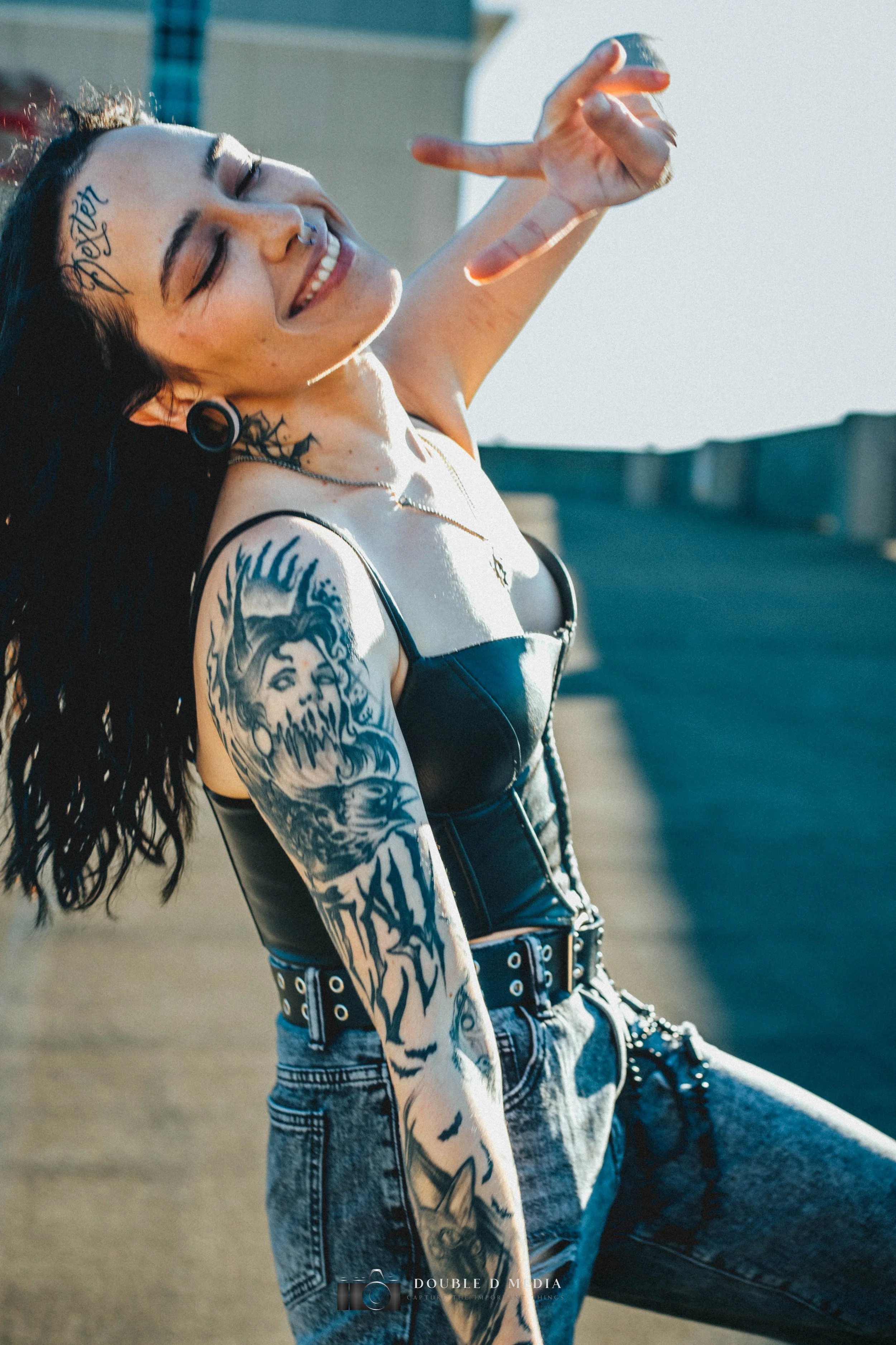 A woman with dark curly hair, tattoos on her arm and face, wearing a black leather top, denim jeans, and black boots, sitting on a rooftop with a cityscape in the background, smiling, and making a peace sign with her hand.