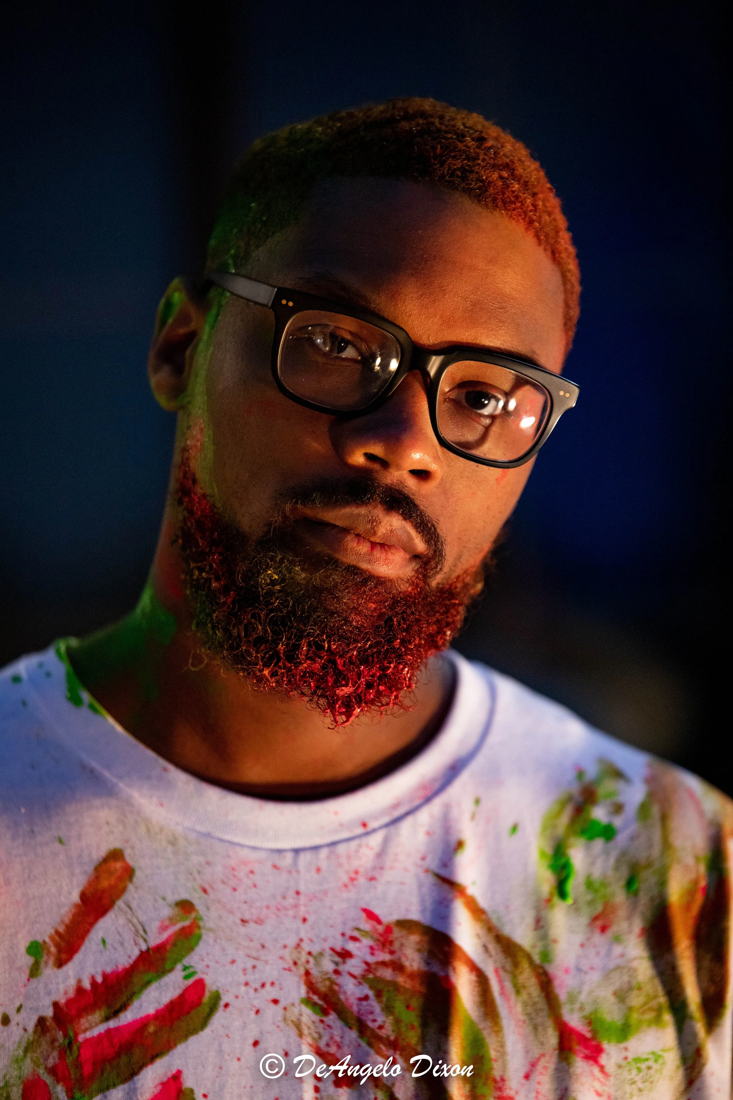 A young man with a reddish-brown beard wearing black glasses. His face has colorful paint splatters in green, red, and yellow. He is wearing a white t-shirt with similar paint splatters. The background is dark and out of focus.