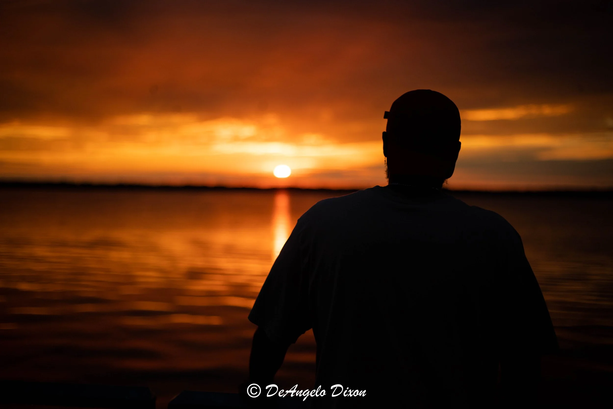 Person with short hair and glasses, in silhouette, watching a sunset over a body of water.
