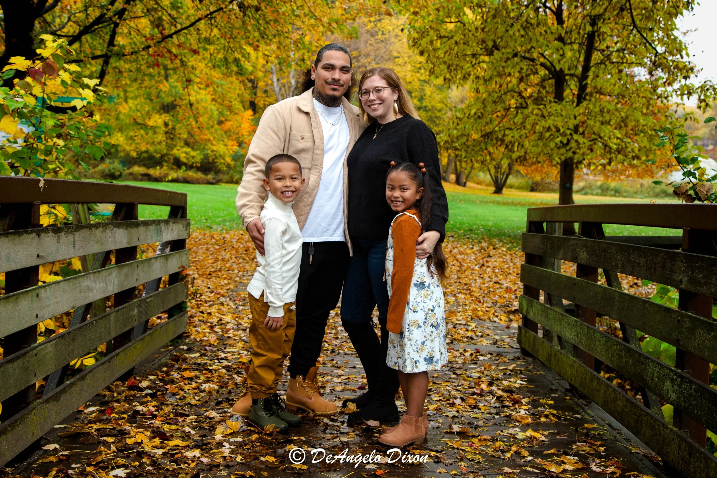 A diverse family of four standing on a bridge in a park during autumn, surrounded by colorful fall foliage; two adults and two children smiling at the camera.