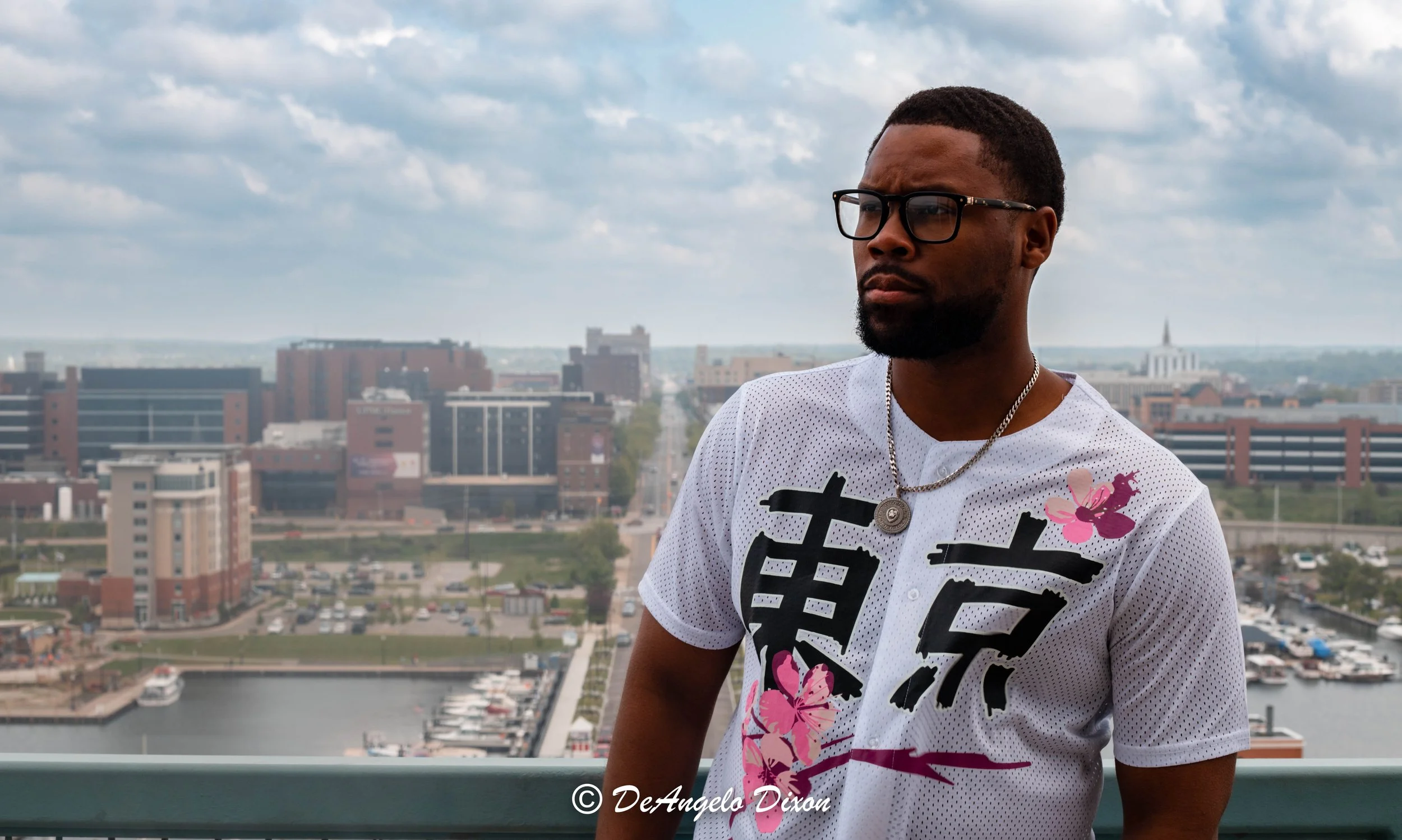 A man wearing glasses, a white shirt with pink flowers and black Kanji characters, and a silver chain with a round pendant, standing on a rooftop with a cityscape and cloudy sky in the background.