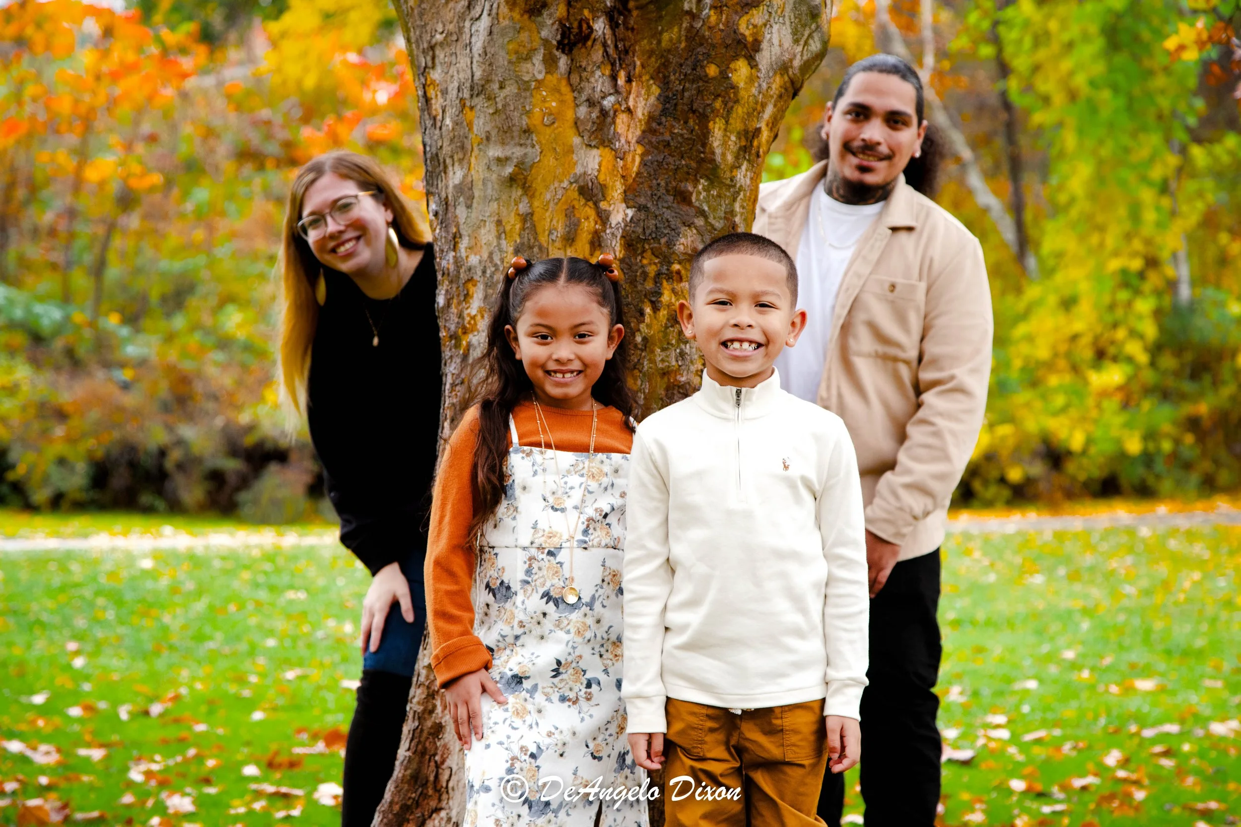 A diverse group of four people, two adults and two children, standing outdoors around a tree with autumn leaves in the background. They are smiling and posing for a photo.