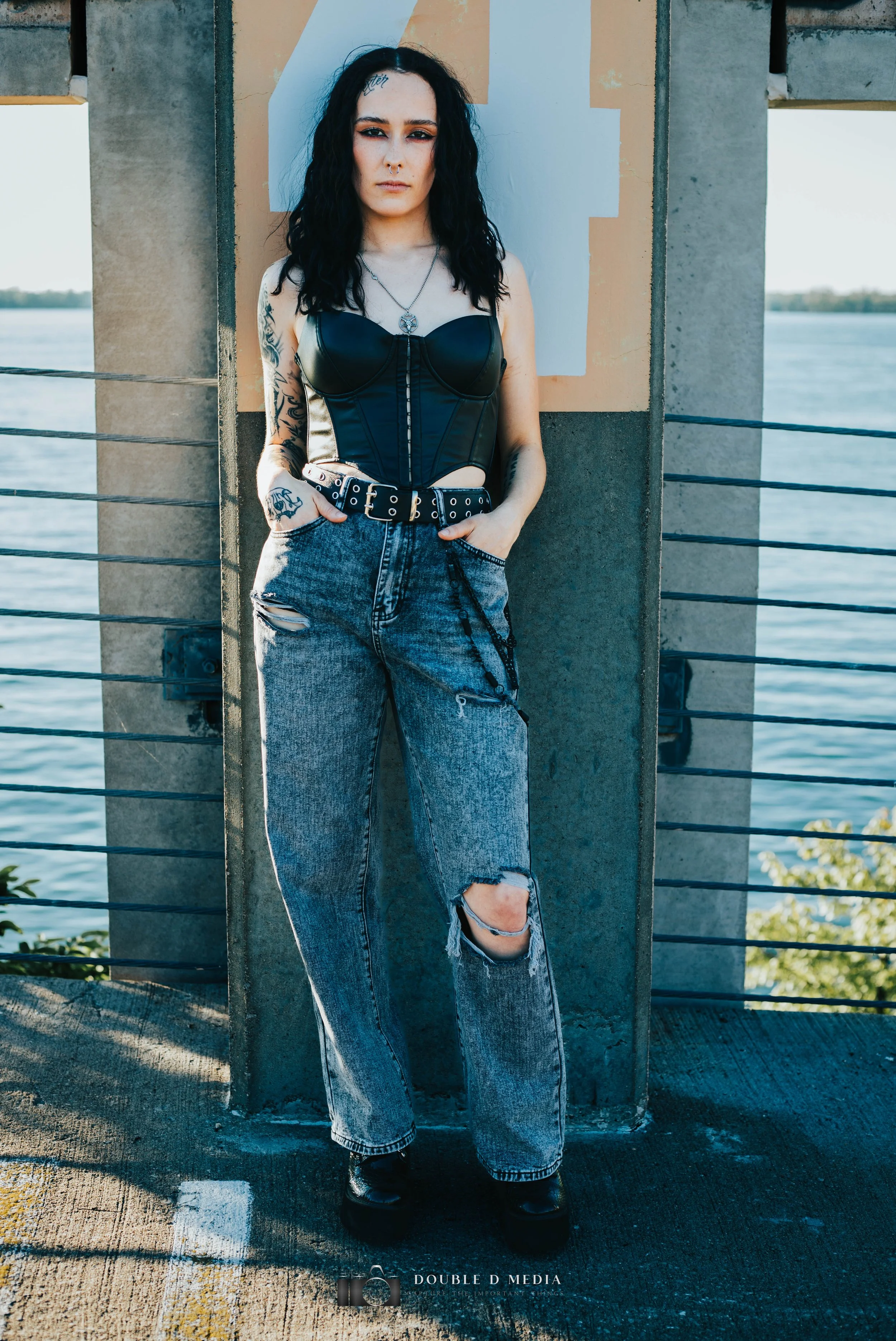 A woman with black curly hair and tattoos on her arms, standing outdoors near a body of water, wearing a black leather corset, ripped jeans, and combat boots.