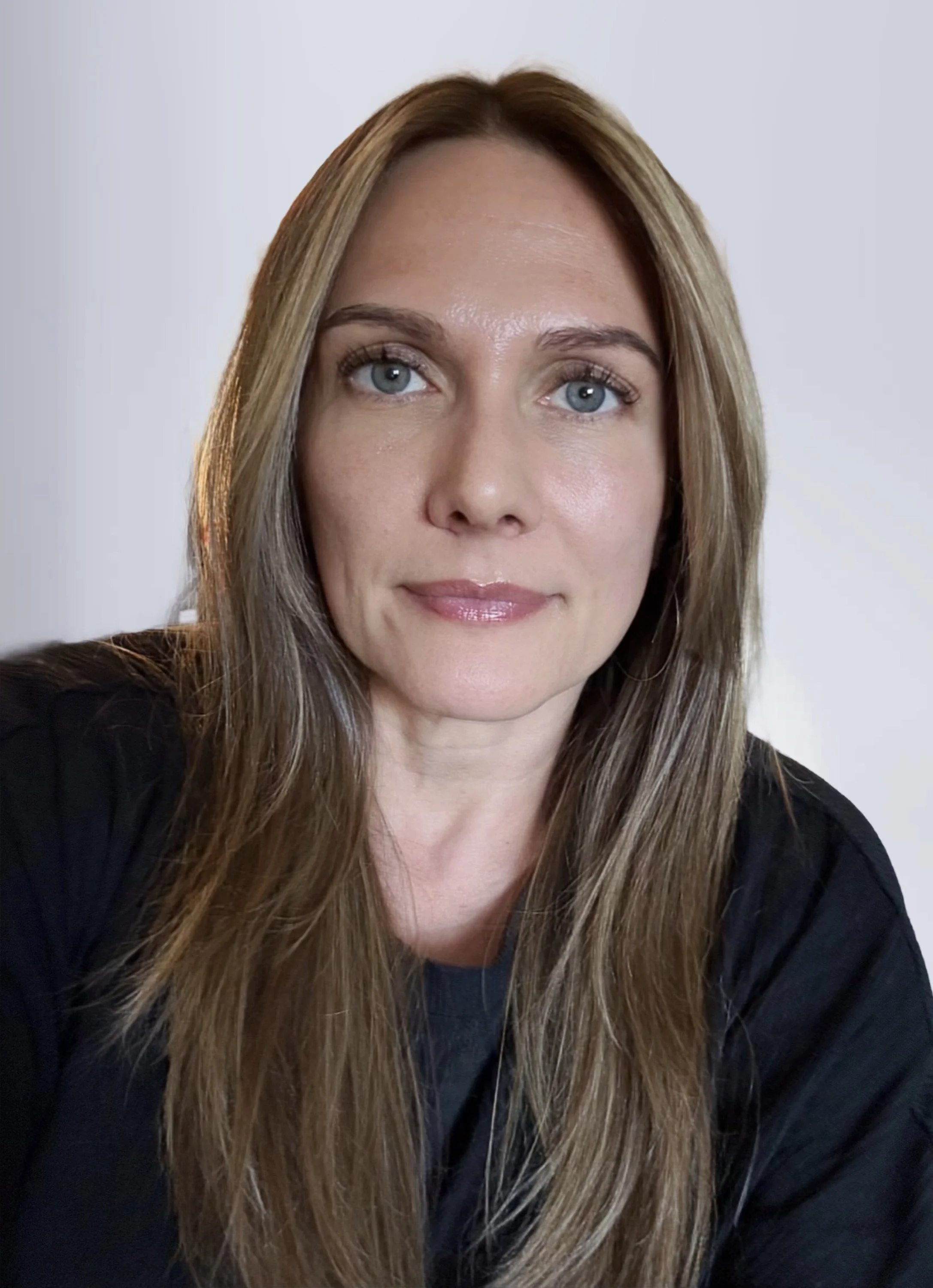 Close-up of a woman with long, light brown hair and blue eyes, smiling slightly, wearing a black top, against a plain light background.