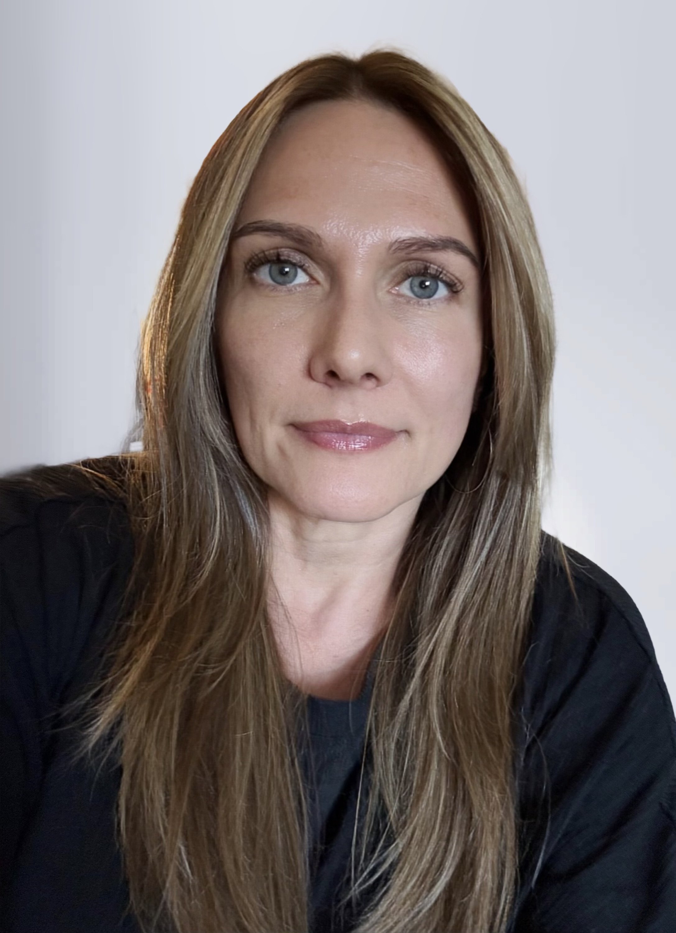 A woman with long, light brown hair and blue eyes, looking into the camera against a plain, light-colored background.
