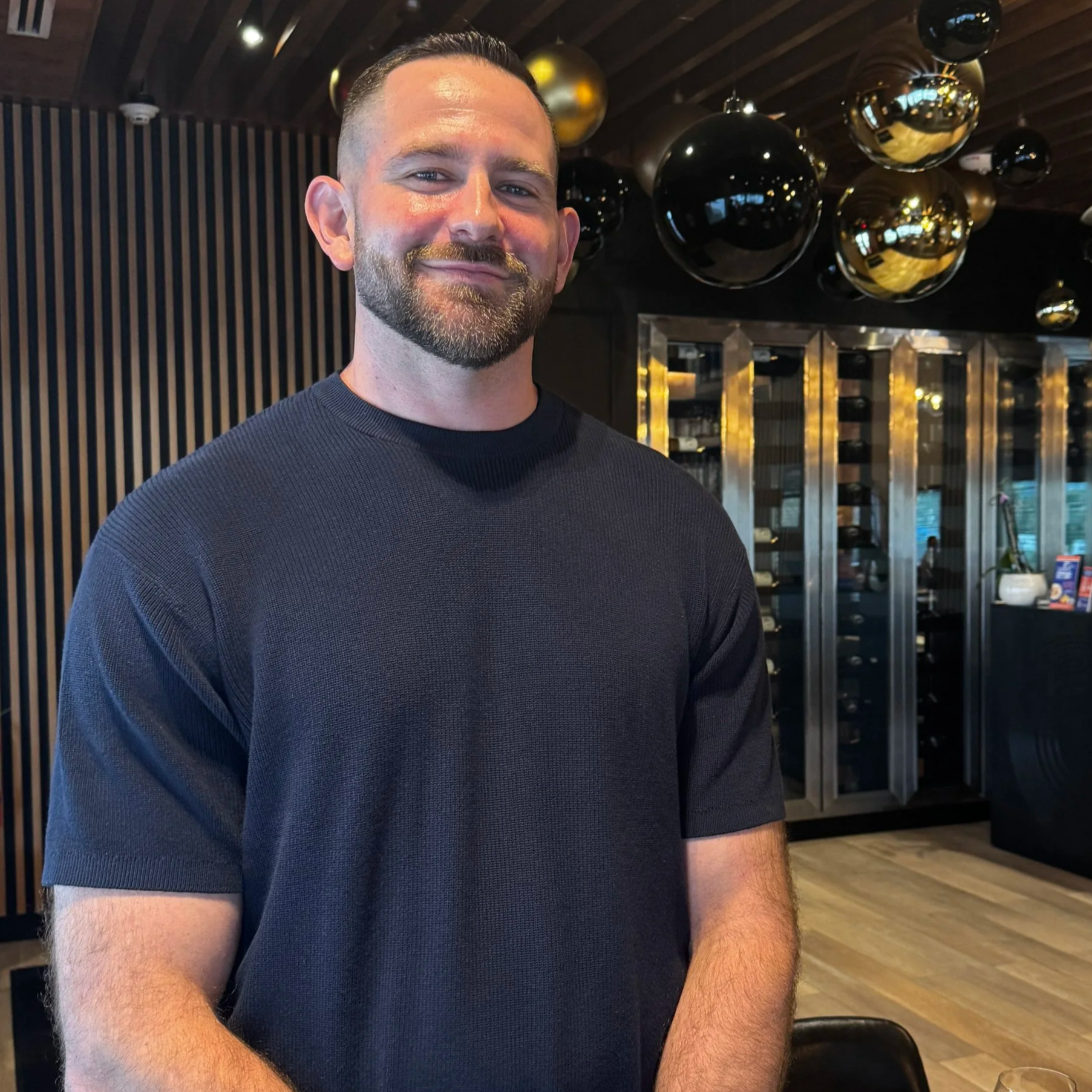 A man with short dark hair, a beard, and light skin, smiling softly, wearing a dark shirt, standing indoors with decorative black and gold balloons hanging from the wooden ceiling, and a modern wine cooler or display case in the background.