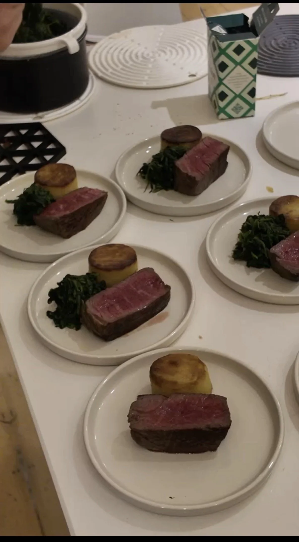 Several white plates with raw steak, roasted potatoes, and sautéed greens on a white countertop. In the background, there are black and white coasters, a paper carton, and some kitchen utensils.