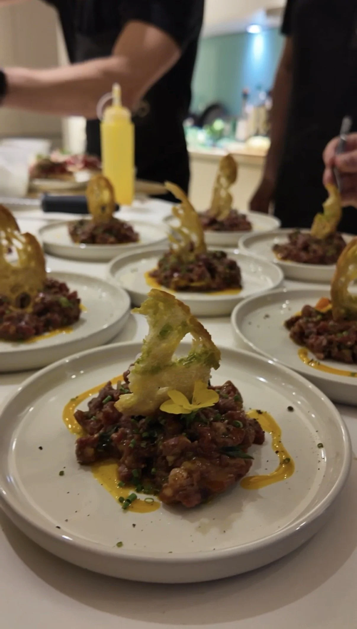 Close-up of plates with beef tartare topped with cheese crisps and garnished with chives and edible flowers, drizzled with yellow sauce, in a restaurant setting.