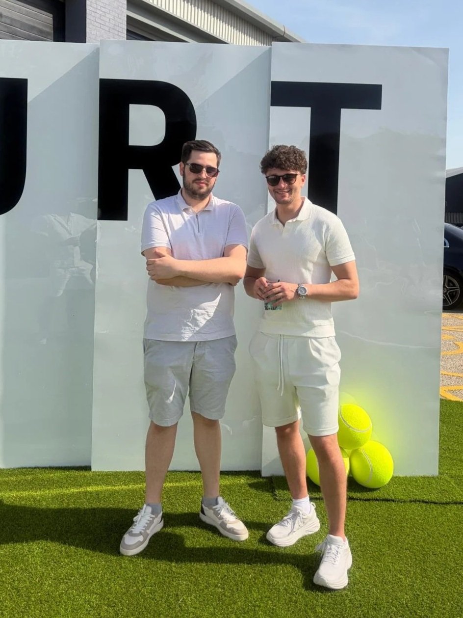 Two young men standing outdoors on a green grassy area in front of a large white panel with the letters 'JRT' on it. Both are wearing sunglasses, white shirts, and shorts, smiling at the camera. There are three tennis balls glowing yellow at their feet.