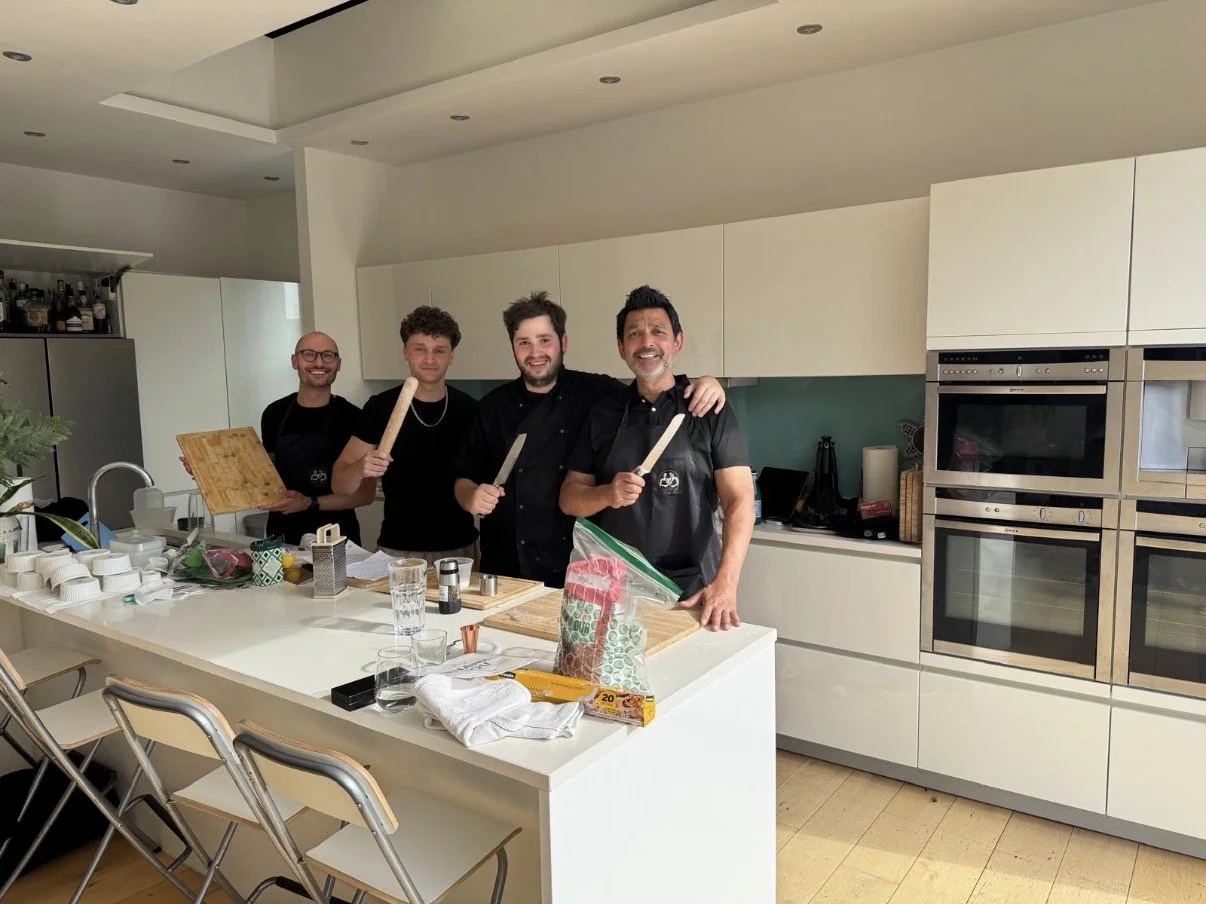 Four men in black aprons standing behind a kitchen island, smiling and holding kitchen utensils, in a modern kitchen.