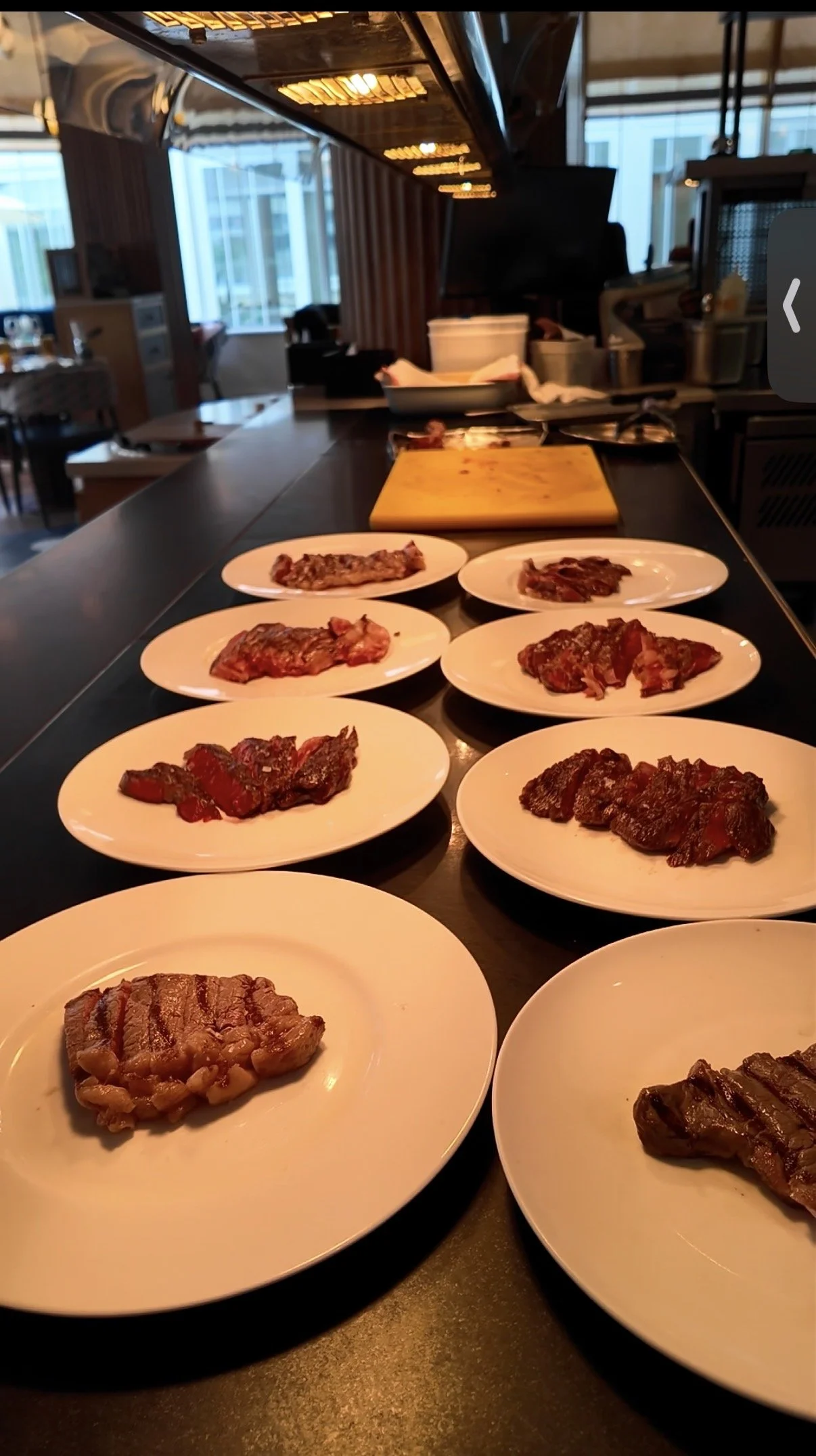 Several plates of raw meat are arranged on a black countertop in a restaurant kitchen, with a wooden cutting board and kitchen equipment in the background.