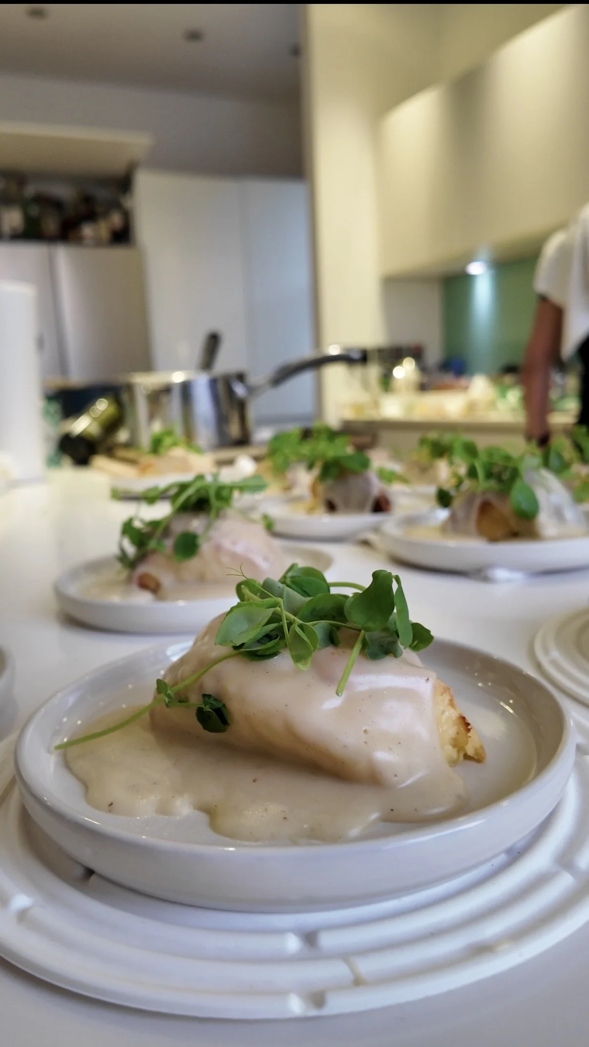 Close-up of individual servings of chicken topped with microgreens on a white plate, with a blurred kitchen background.