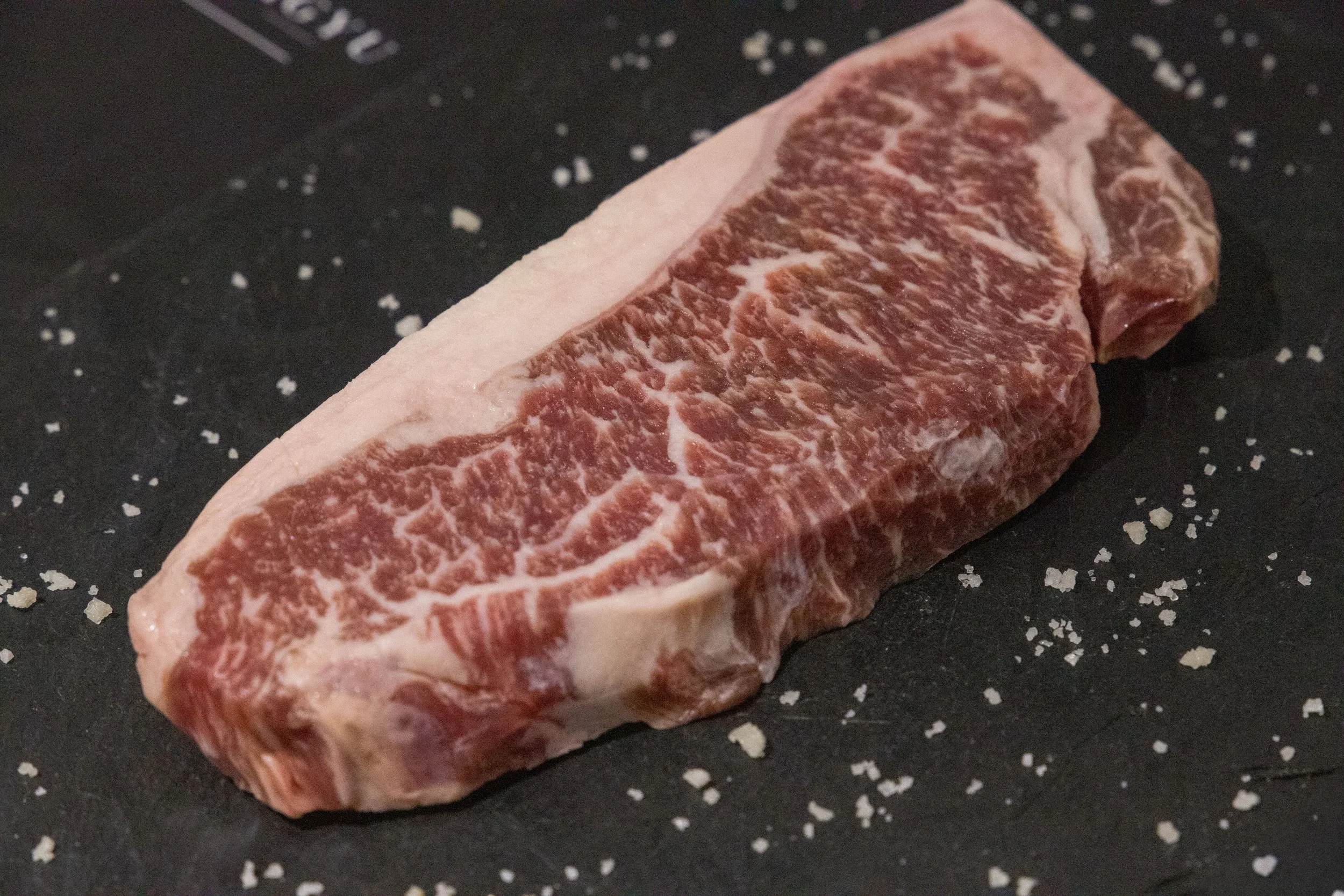 A raw, marbled cut of beef steak on a black surface sprinkled with coarse salt.