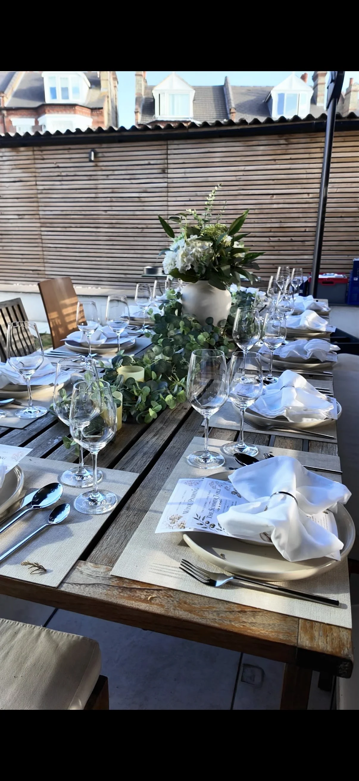 A long wooden outdoor dining table set for a formal event with multiple wine glasses, silverware, and white napkins. A large white vase with a floral arrangement and greenery runs down the center of the table. The setting is in a backyard with a wood