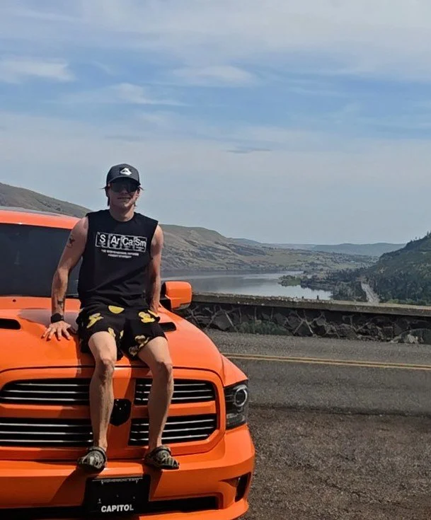 A young man sitting on the hood of an orange car by a scenic lake and hills, wearing a black tank top, black shorts with yellow patterns, sunglasses, and a cap.