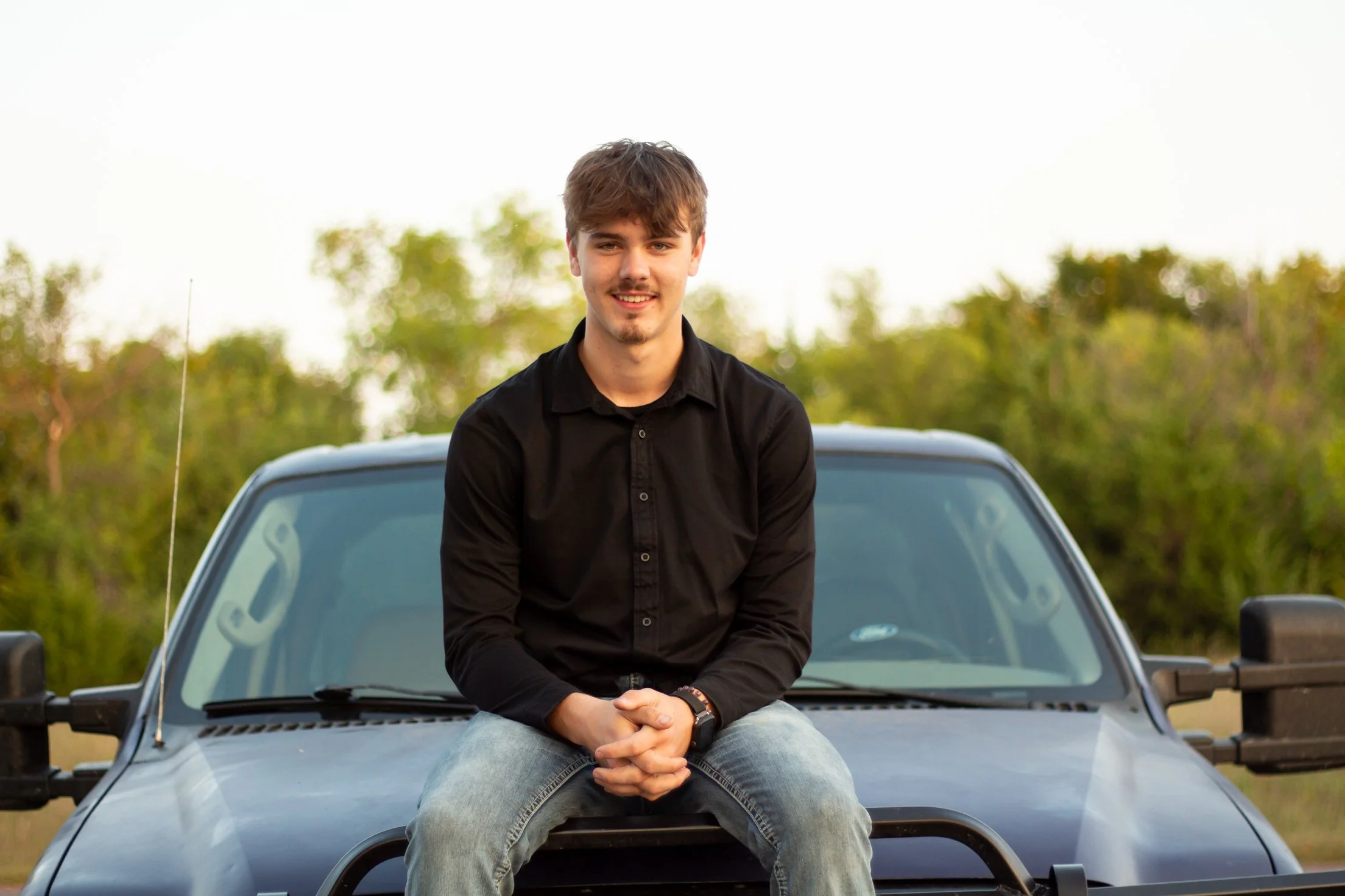 A young man with brown hair, a light beard, and mustache, wearing a black shirt and jeans, sitting on the hood of a gray pickup truck outdoors during daytime. The background features trees and a clear sky.