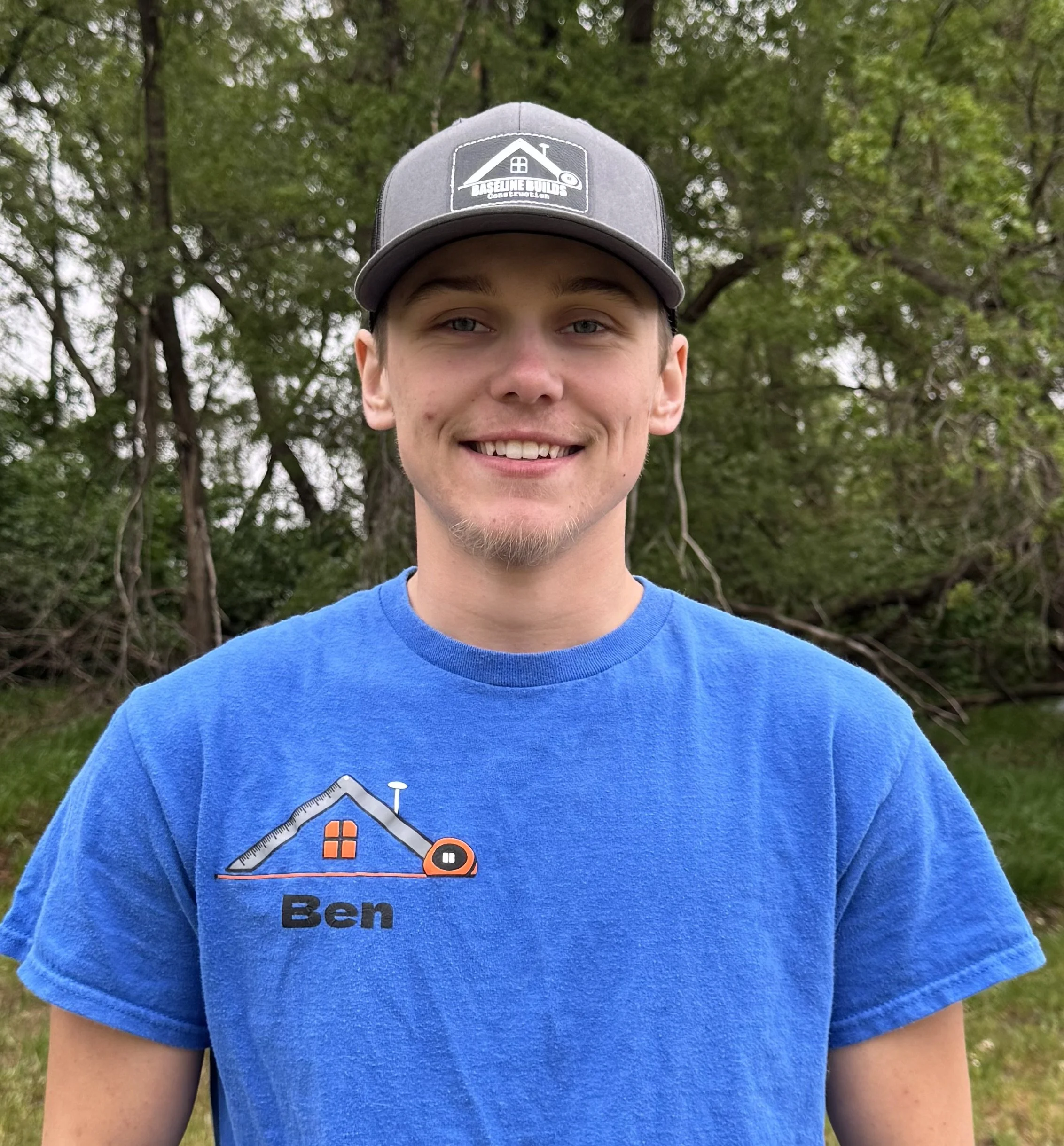 Close-up of a young man with light skin and blue eyes wearing a gray cap with a logo and text, and a blue T-shirt with a house graphic and the name 'Ben' printed on it, standing outdoors in front of trees.