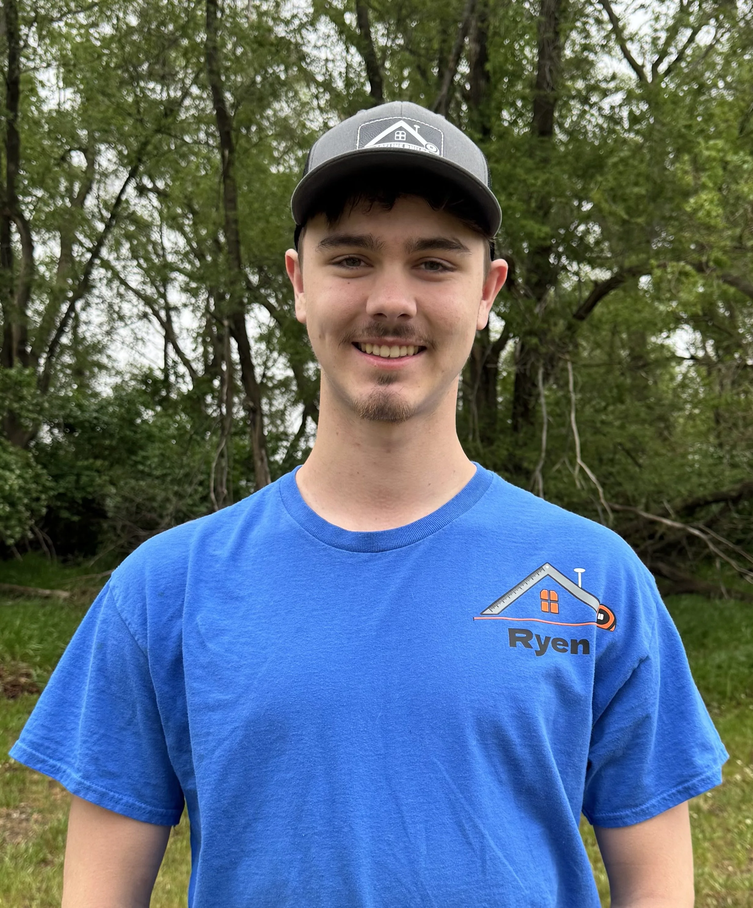 Close-up of a young man outdoors wearing a blue T-shirt with a logo and a gray baseball cap, smiling in front of green trees.