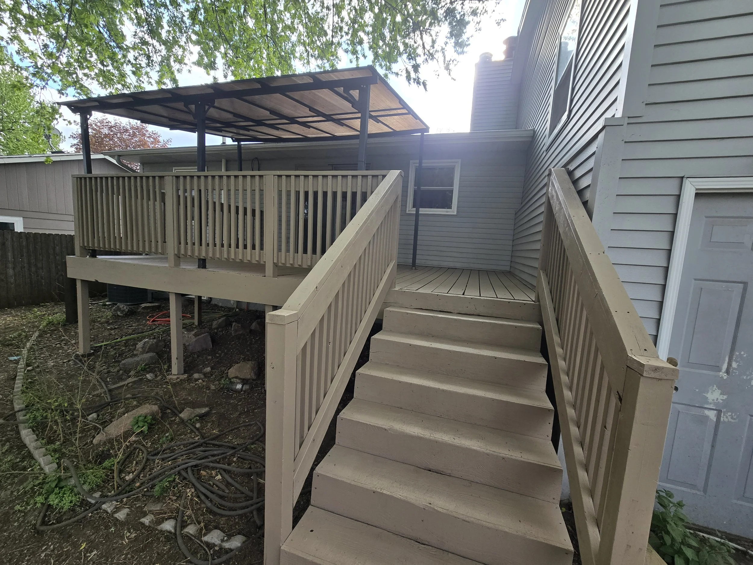 Wooden deck with stairs attached to a house, partially shaded with a roof canopy, yard with soil, rocks, and plants, and trees overhead.