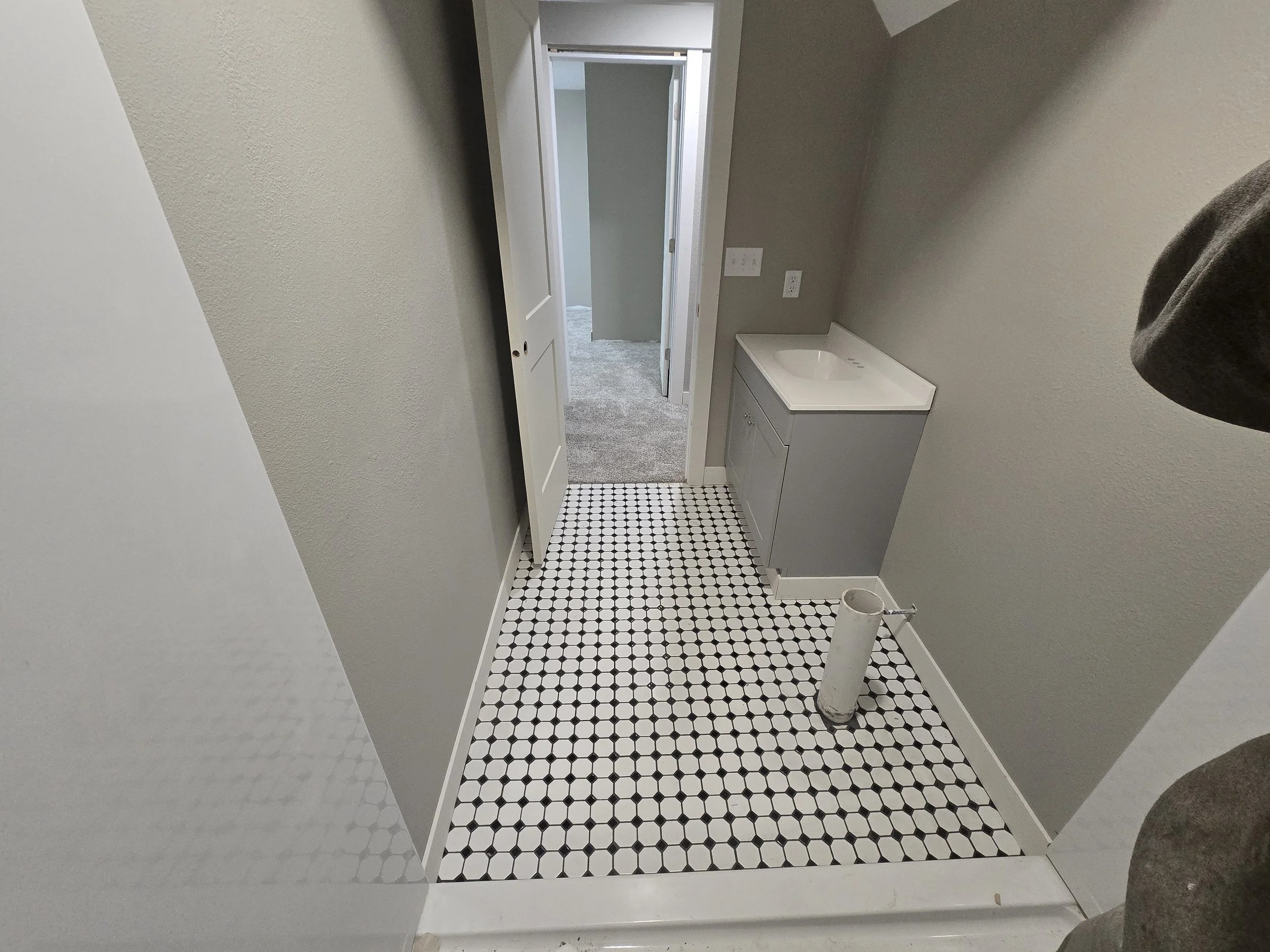 The image shows a small bathroom with a black and white hexagonal tile floor, a gray vanity with a sink, a white pipe visible in the corner, and an open doorway leading to a carpeted room with light-colored walls and a closet with a door.