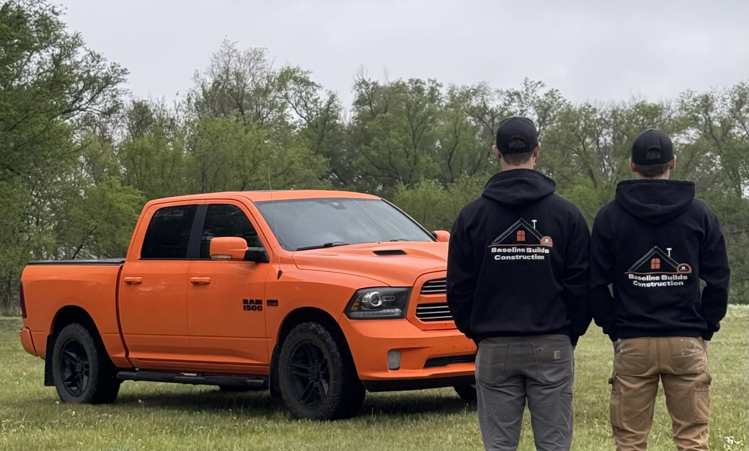 Two men wearing black jackets with a house logo and the words 'Baseline Builds Construction' on the back stand outdoors, facing away from the camera, near an orange pickup truck during daytime with overcast sky and green trees in the background.