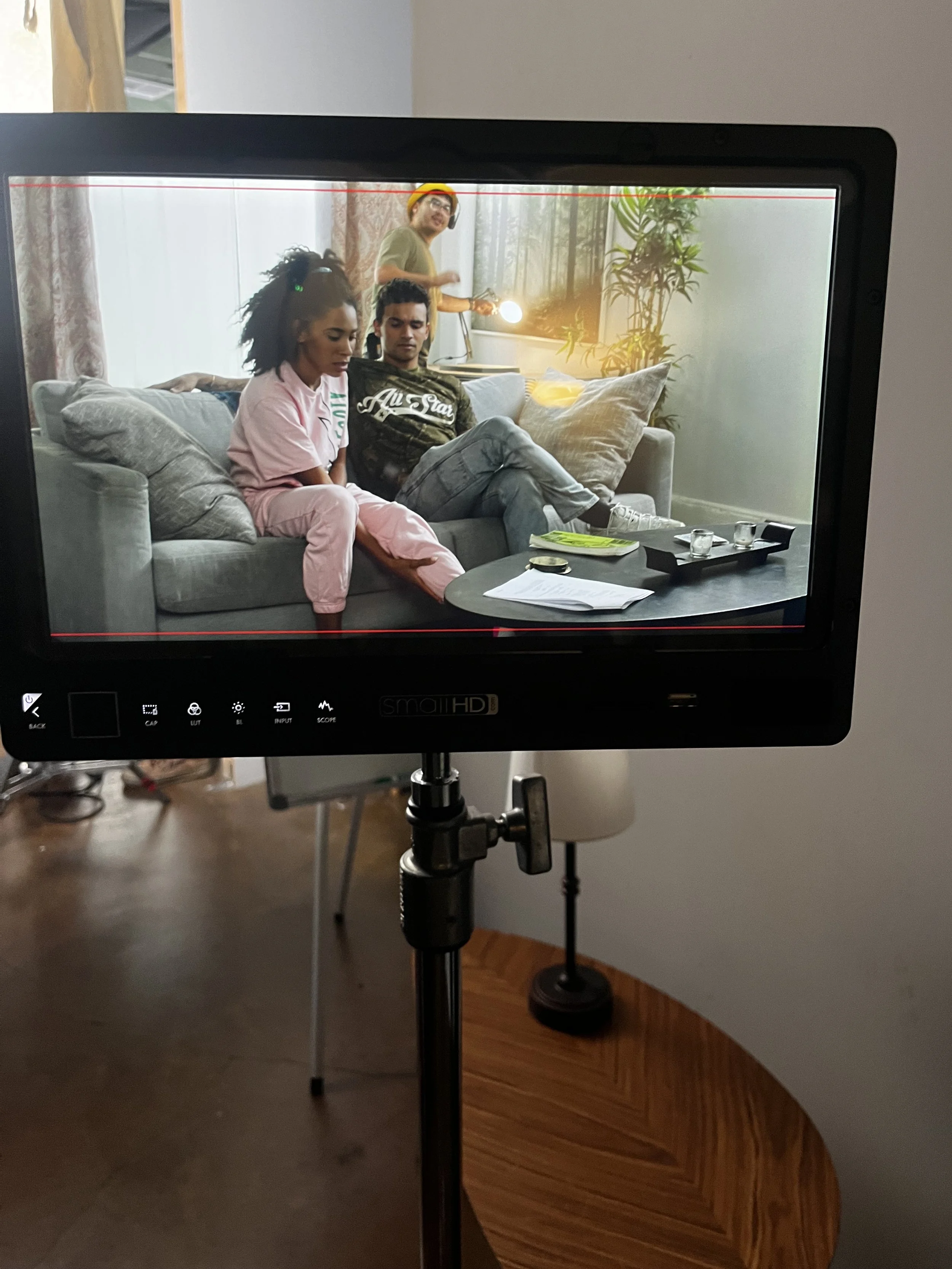 A camera monitor displays a scene of three people in a living room, with two sitting on a couch and one standing behind them, while recording.