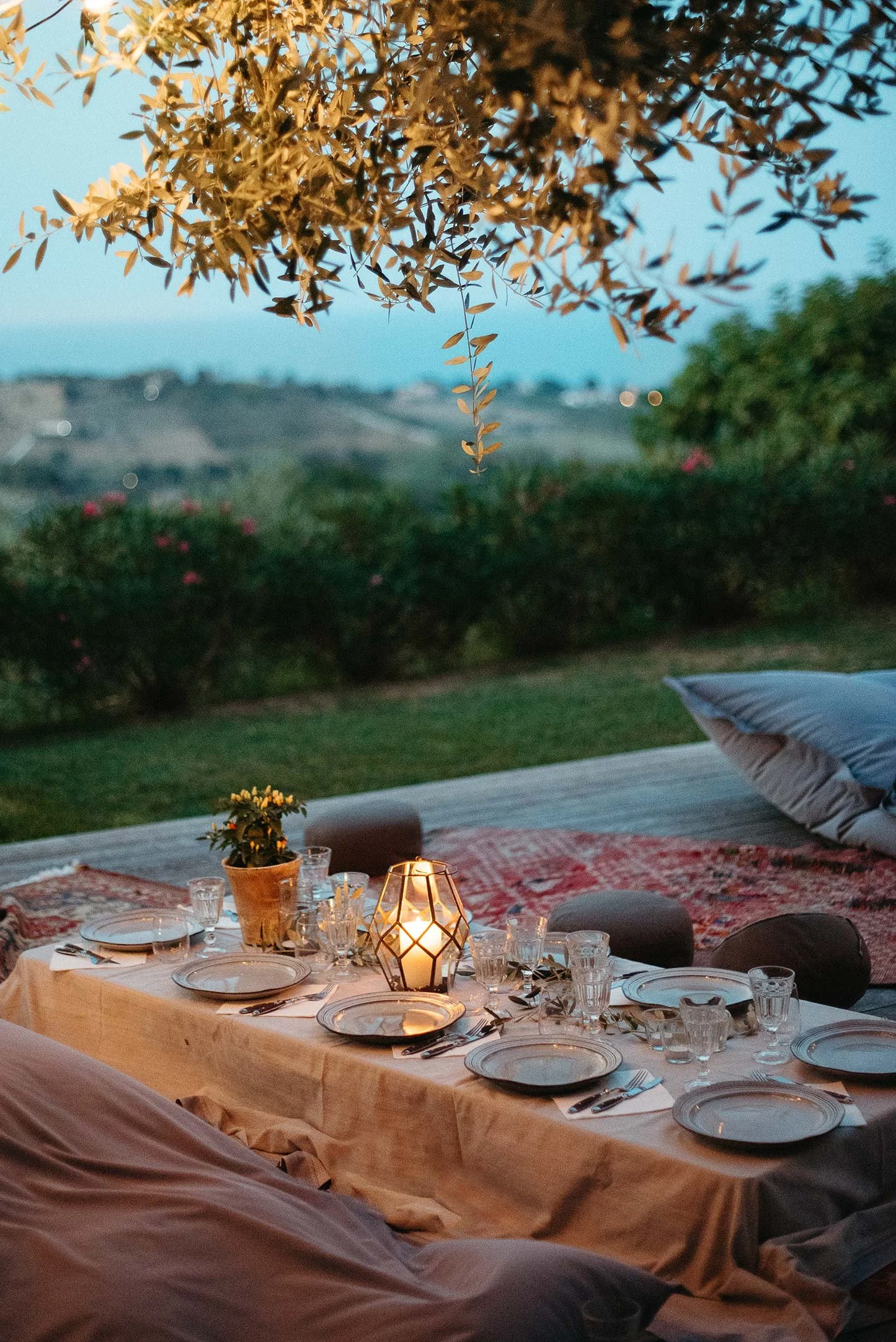 A cozy outdoor dining setting with a long table covered with a white tablecloth, set with plates, glasses, cutlery, and a central candle lantern. There are cushions on the ground for seating. The background shows a grassy area with trees and hills under a twilight sky, creating a warm and inviting atmosphere for an evening gathering.