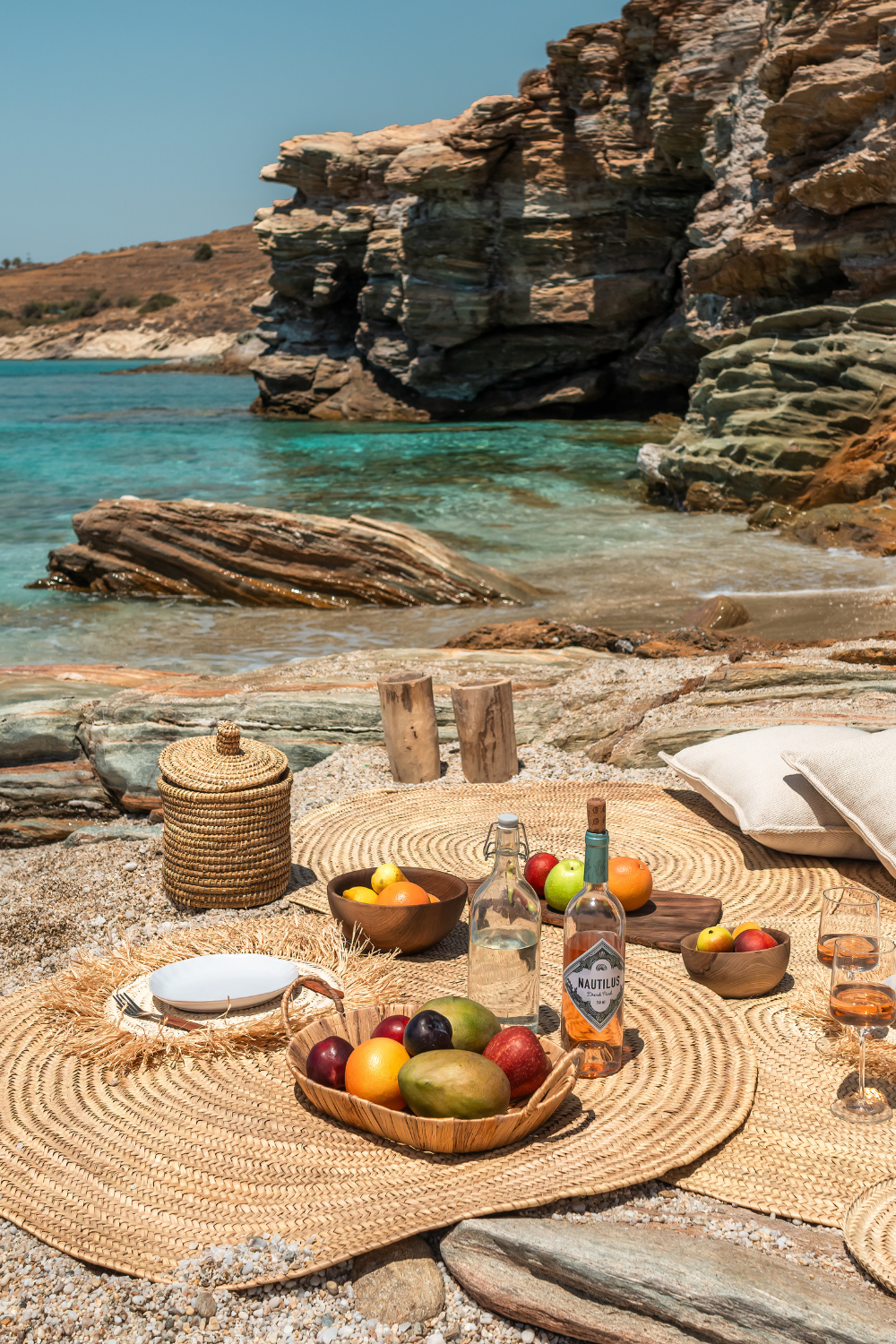Beachside picnic setup with woven mats, bowls of fruit, bottles of water and wine, glasses, and pillows on rocks near the ocean with rocky cliffs in the background.