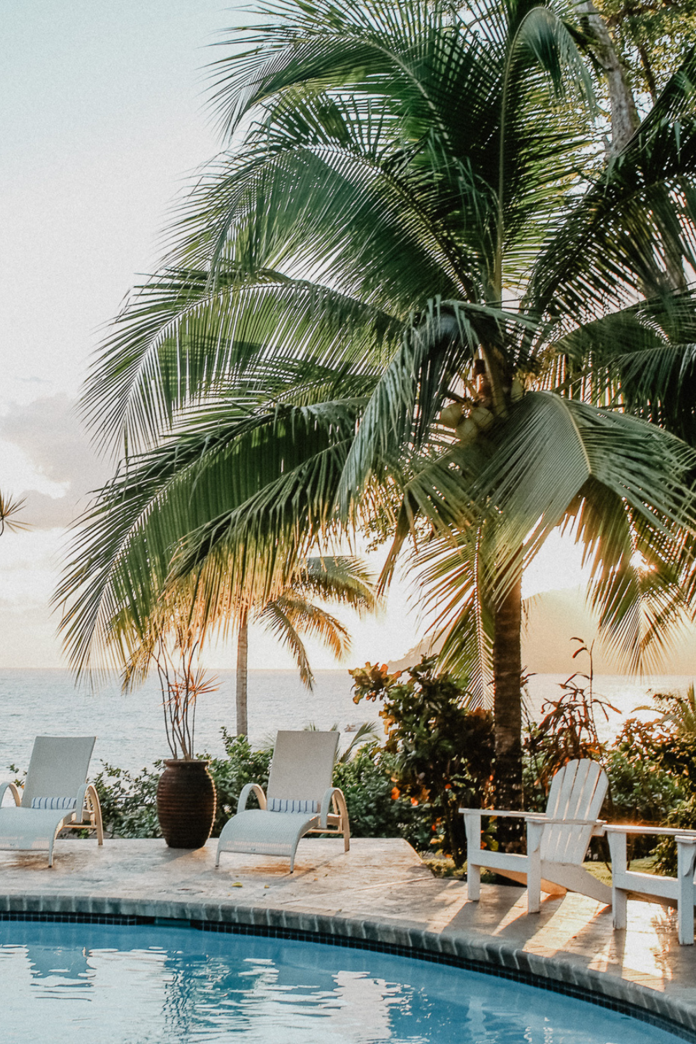 Tropical poolside scene with palm trees, lounge chairs, and the ocean in the background during sunset.
