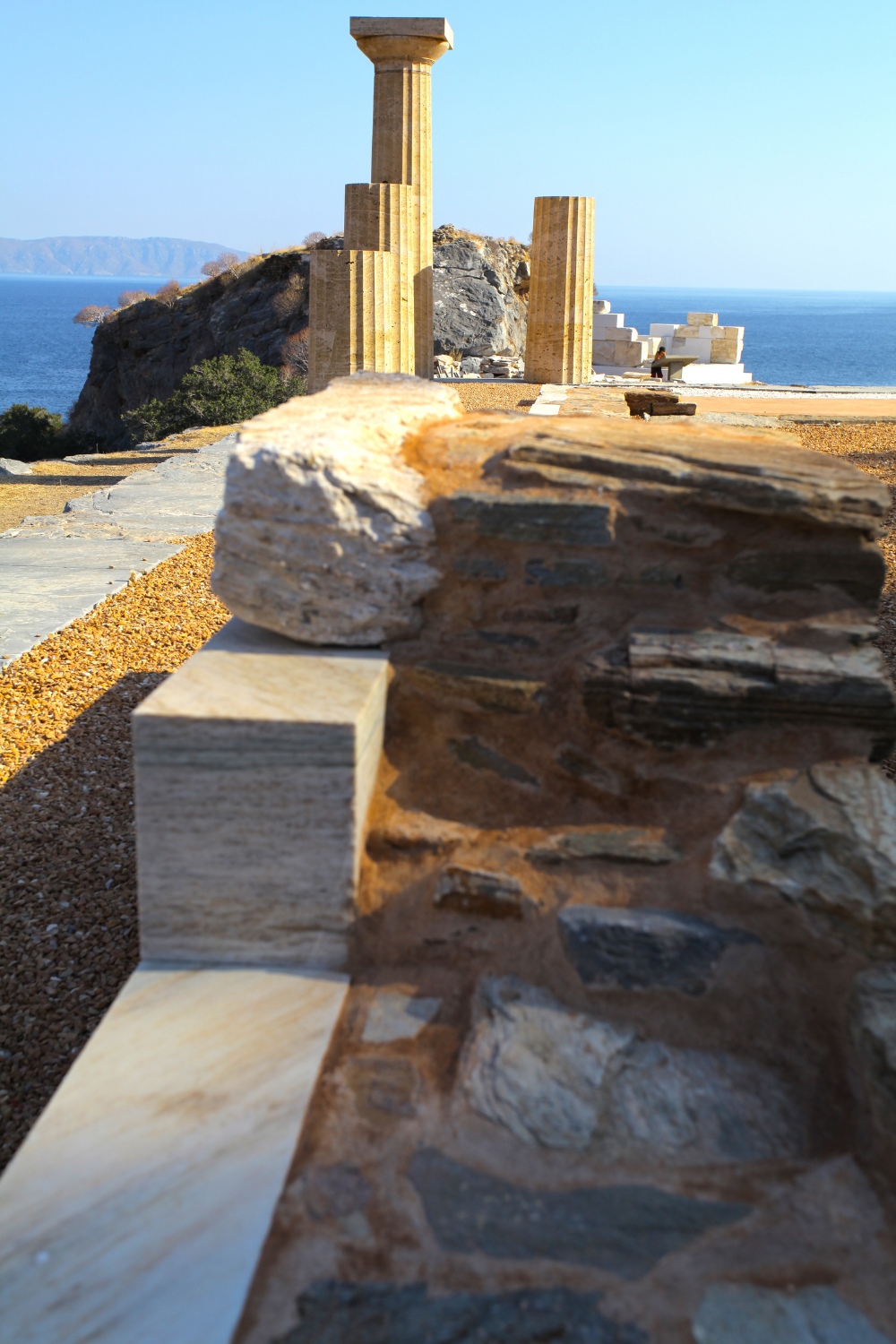 Ancient ruins with stone columns and rocks overlooking the sea, with a person in the background sitting on steps by the water.