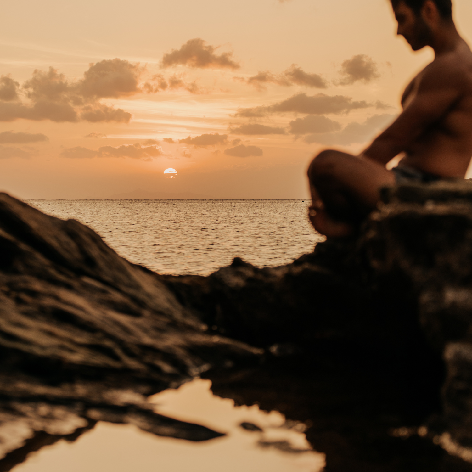 A person sitting on rocks by the ocean at sunset,