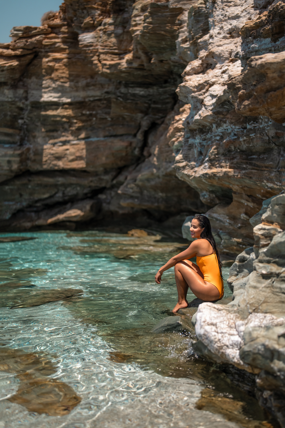A woman in an orange swimsuit sitting on rocks by a clear, turquoise water cove with rocky cliffs in the background.