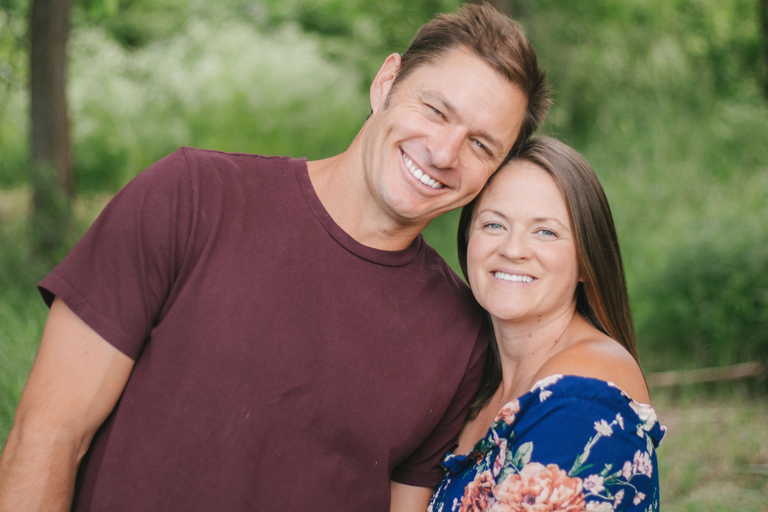 A smiling man and woman standing close together outdoors in a green, wooded area, with the man wearing a maroon t-shirt and the woman wearing a blue floral dress.
