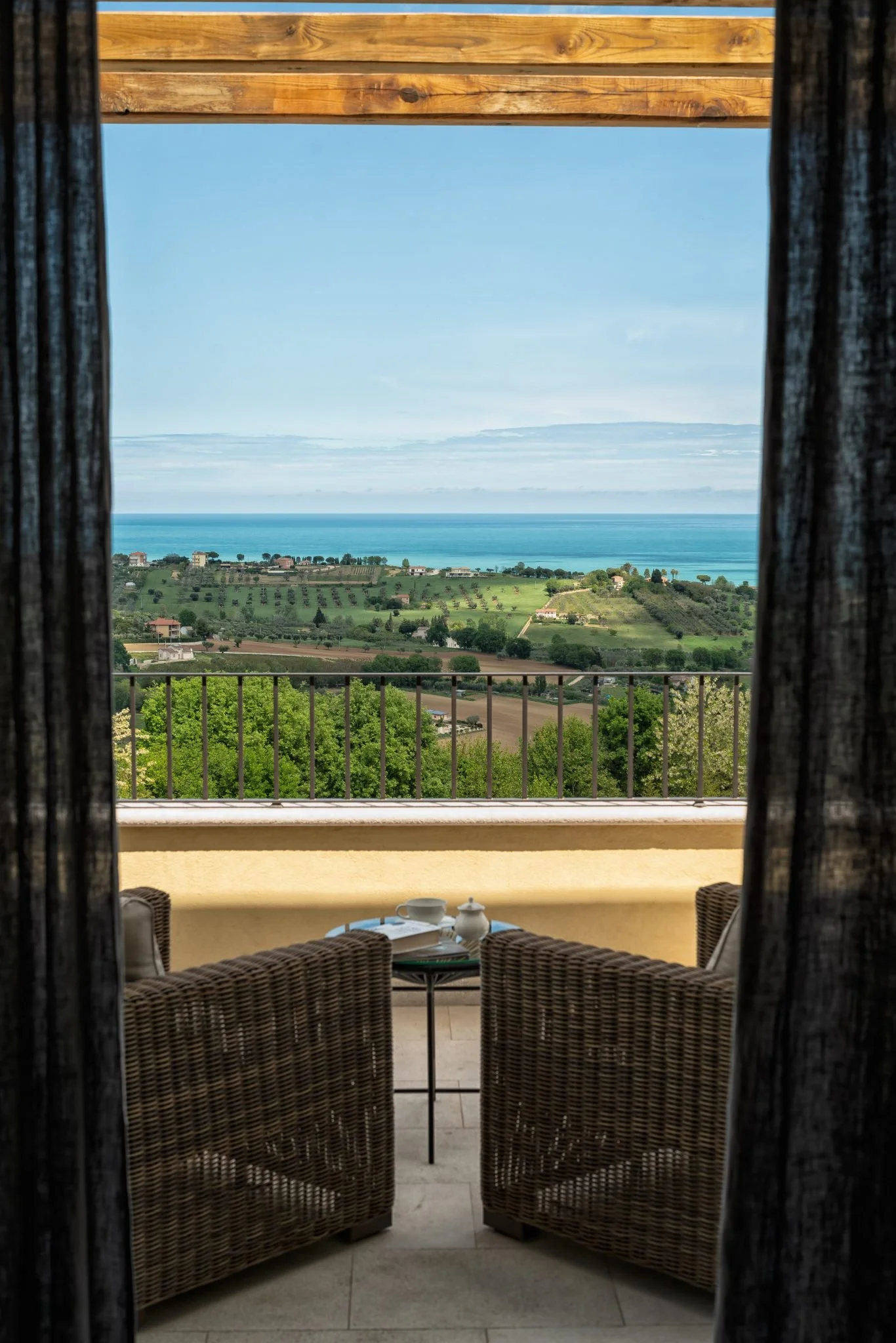 View from a balcony showing wicker chairs and a small table with a teapot and cups, overlooking rolling green hills, vineyards, and the ocean under a partly cloudy sky.