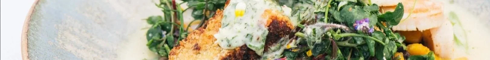 Close-up of a plated dish featuring a breaded and fried piece of fish, topped with a creamy herb sauce, served with a side of fresh greens and vegetables.