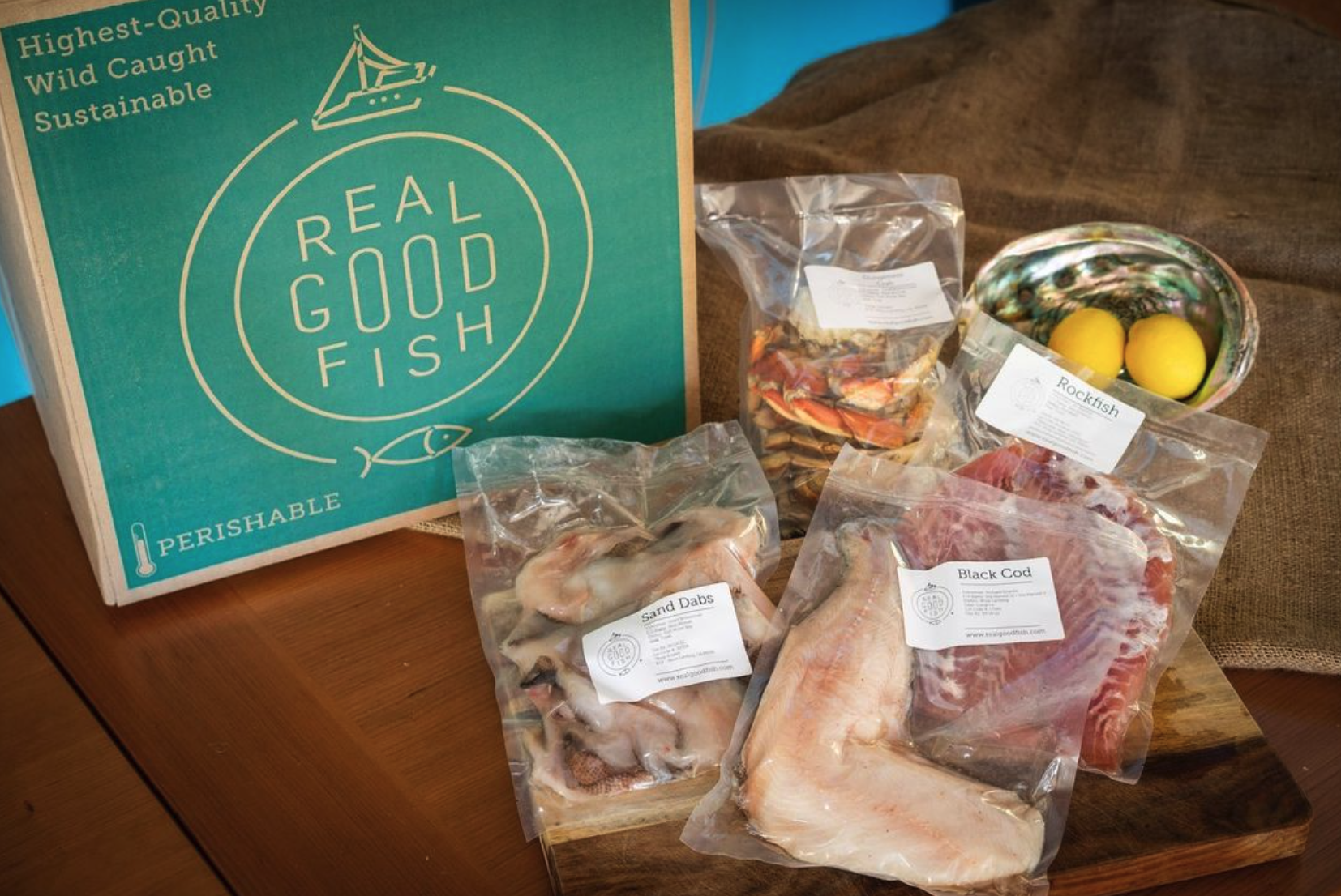 A package of seafood labeled 'Real Good Fish' with various fish and shellfish in plastic bags, including black cod, rockfish, sand dabs, and a bag of crab legs. There is also a colorful bowl with lemons.
