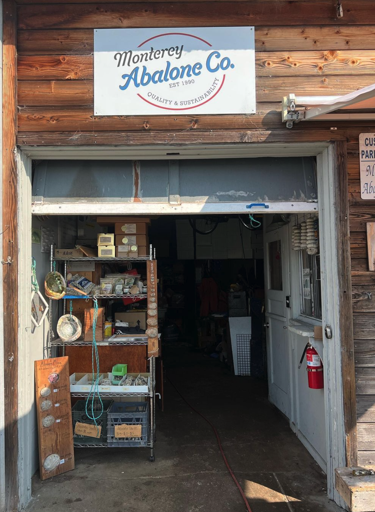 Entrance to a store called Monterey Abalone Co. with a sign indicating it was established in 1990, featuring products like abalone shells and other items on display on shelves and a wooden board outside.