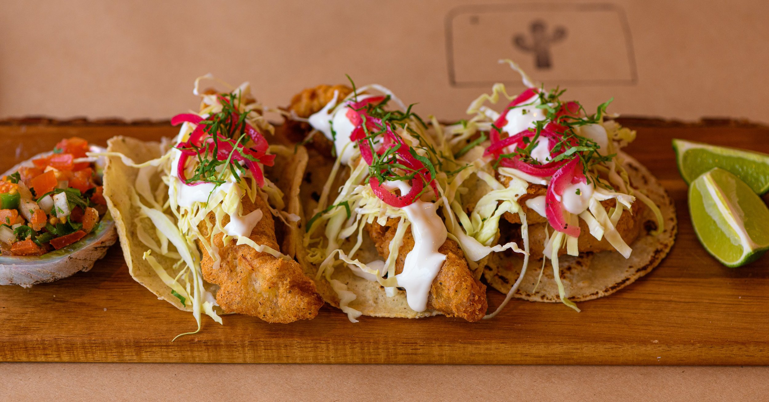 Fish tacos topped with shredded lettuce, sliced red onions, and garnished with chopped herbs, served on a wooden board with lime wedges and a side of diced vegetable salsa.