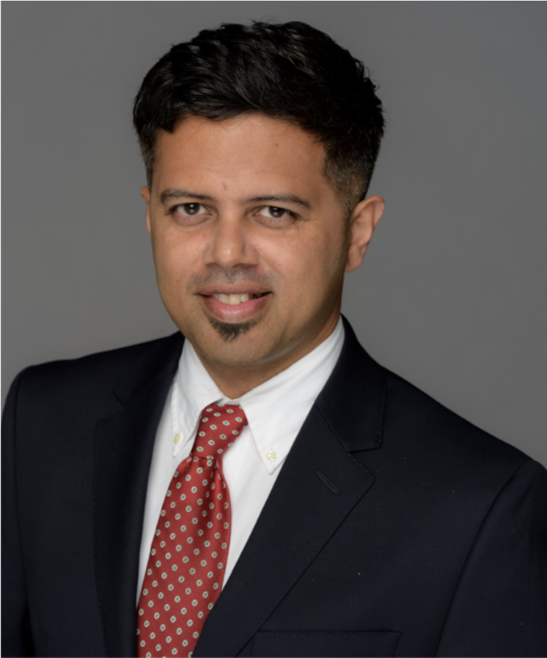 Professional headshot of a man with dark hair, wearing a black suit, white shirt, and red tie with small white and green dots, against a gray background.